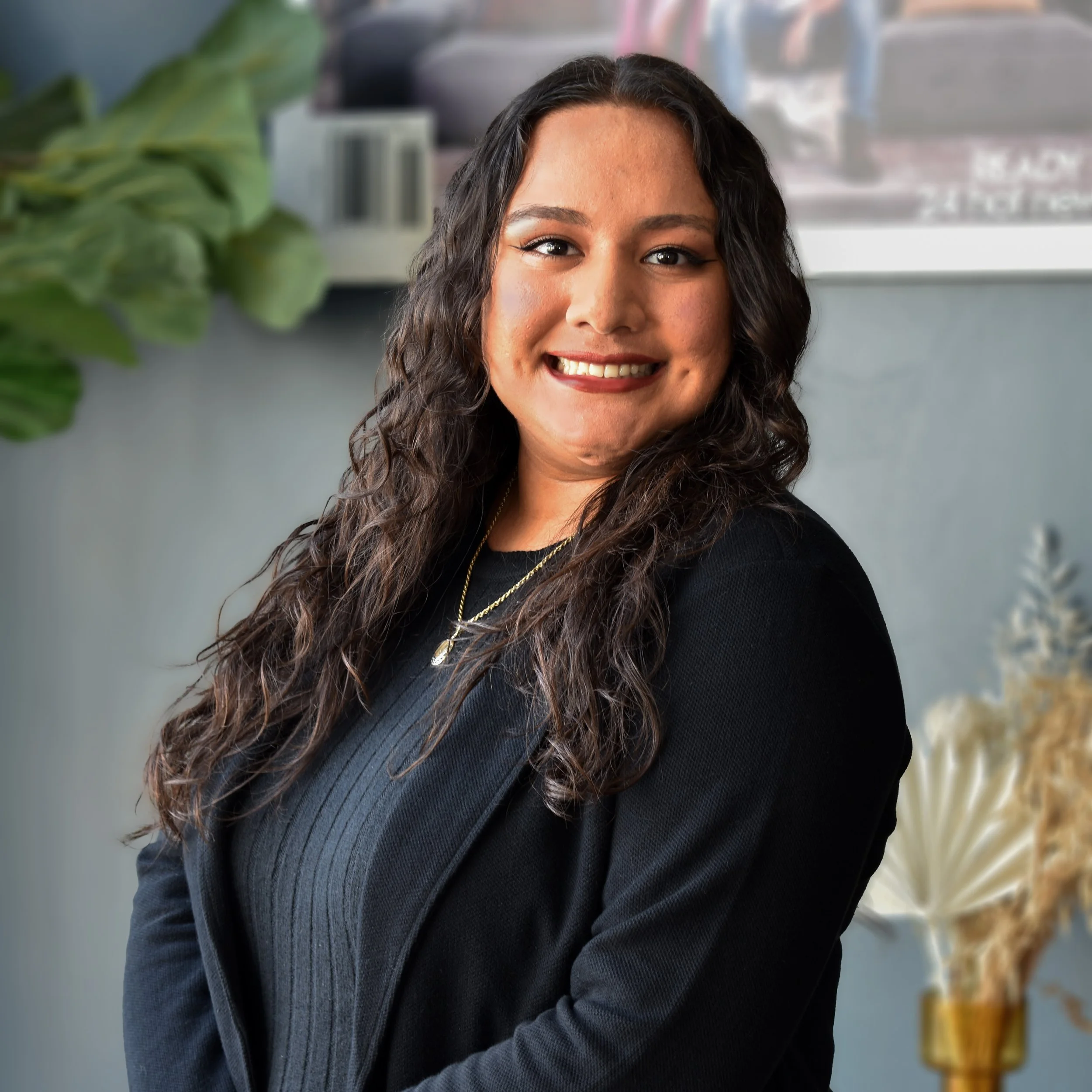 A young woman with long, curly dark hair smiling, wearing a black blazer and layered necklaces, standing in a modern indoor setting with plants and decorative items in the background.