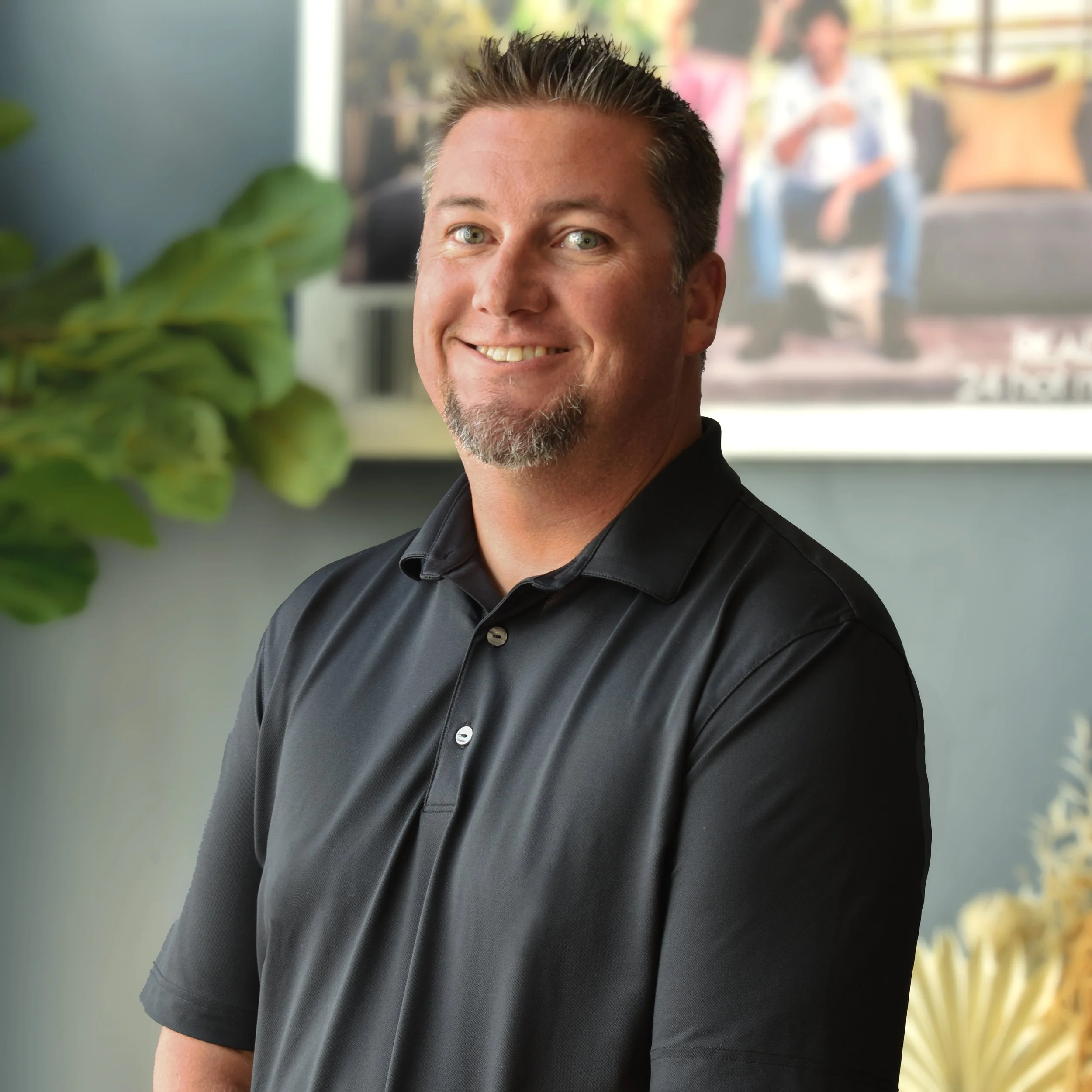 A man with short dark hair and a goatee, wearing a black polo shirt, smiling in front of a blurred background with a plant and a photograph of two people sitting on a couch.