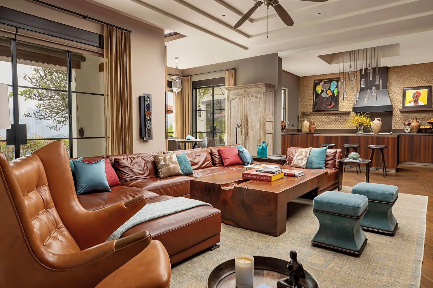 Living room with leather and fabric sofas, a large wooden coffee table, colorful pillows, and a modern open kitchen in the background featuring artwork, vases, and a yellow flowers arrangement.