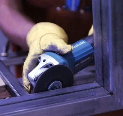 Person using an electric power sander on a wooden frame, wearing yellow work gloves.