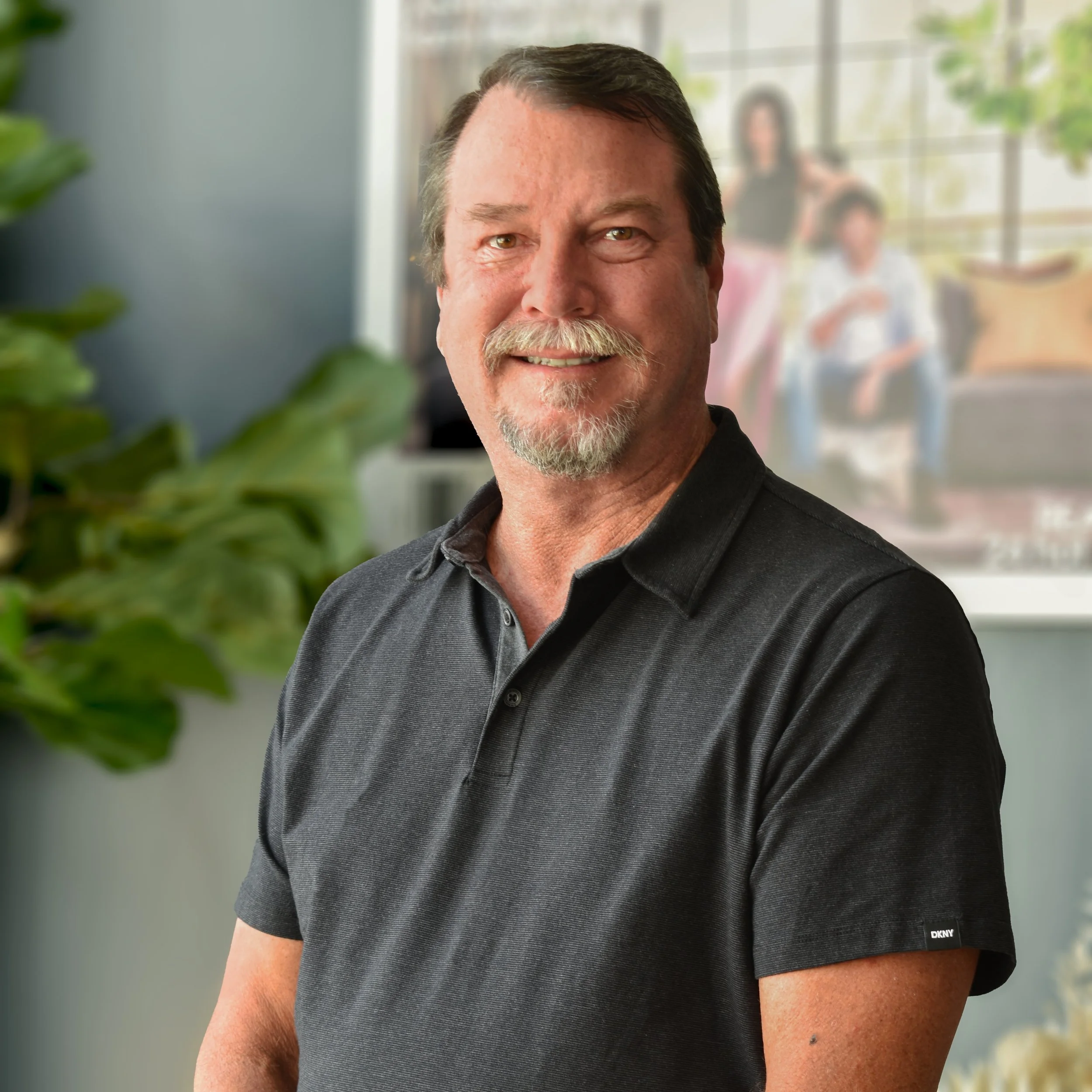 A man with dark hair, a mustache, and a beard smiling, wearing a black polo shirt, standing in a well-lit room with a large window and a blurred family picture in the background.