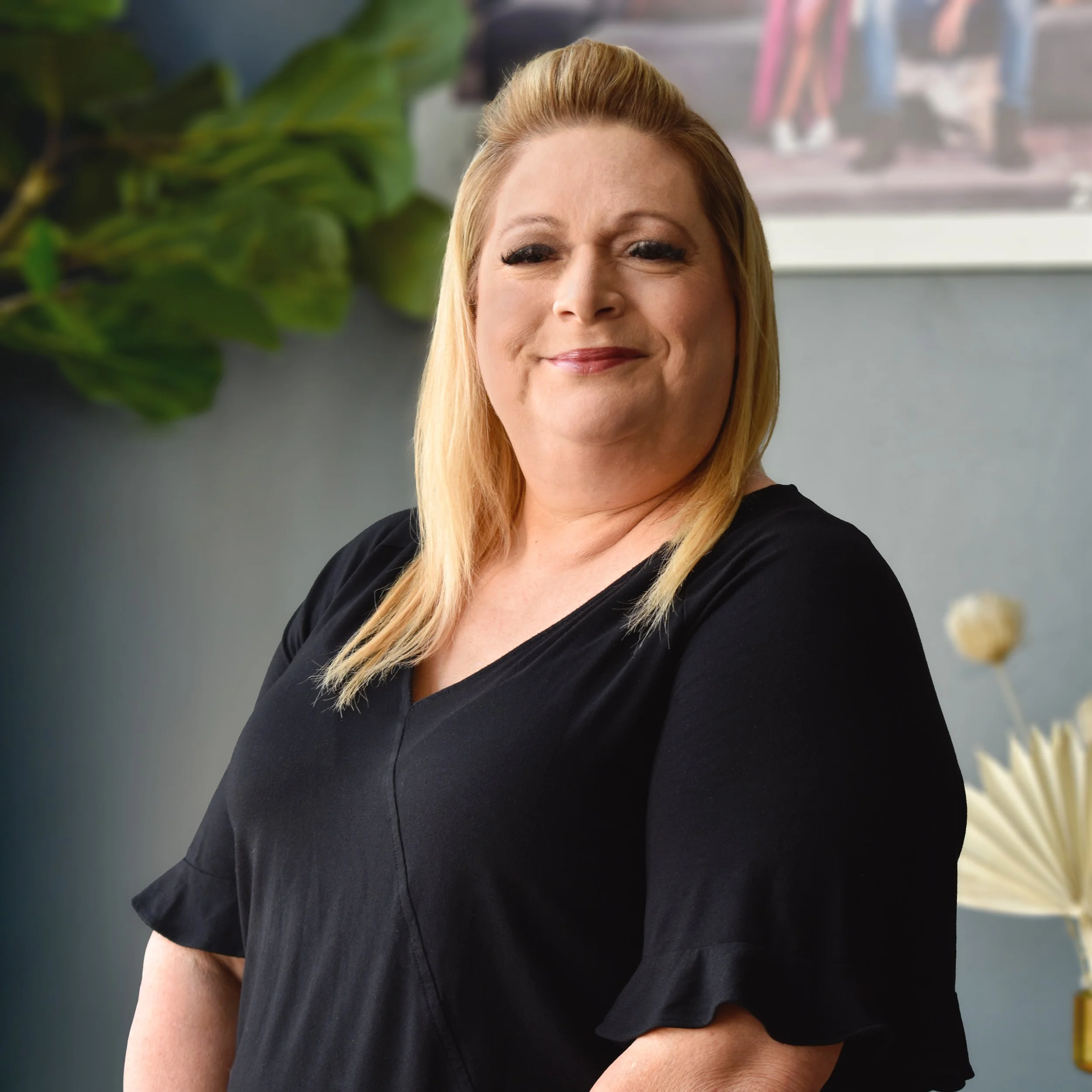 A woman with blonde hair smiling in an indoor setting with green plants and artwork in the background.