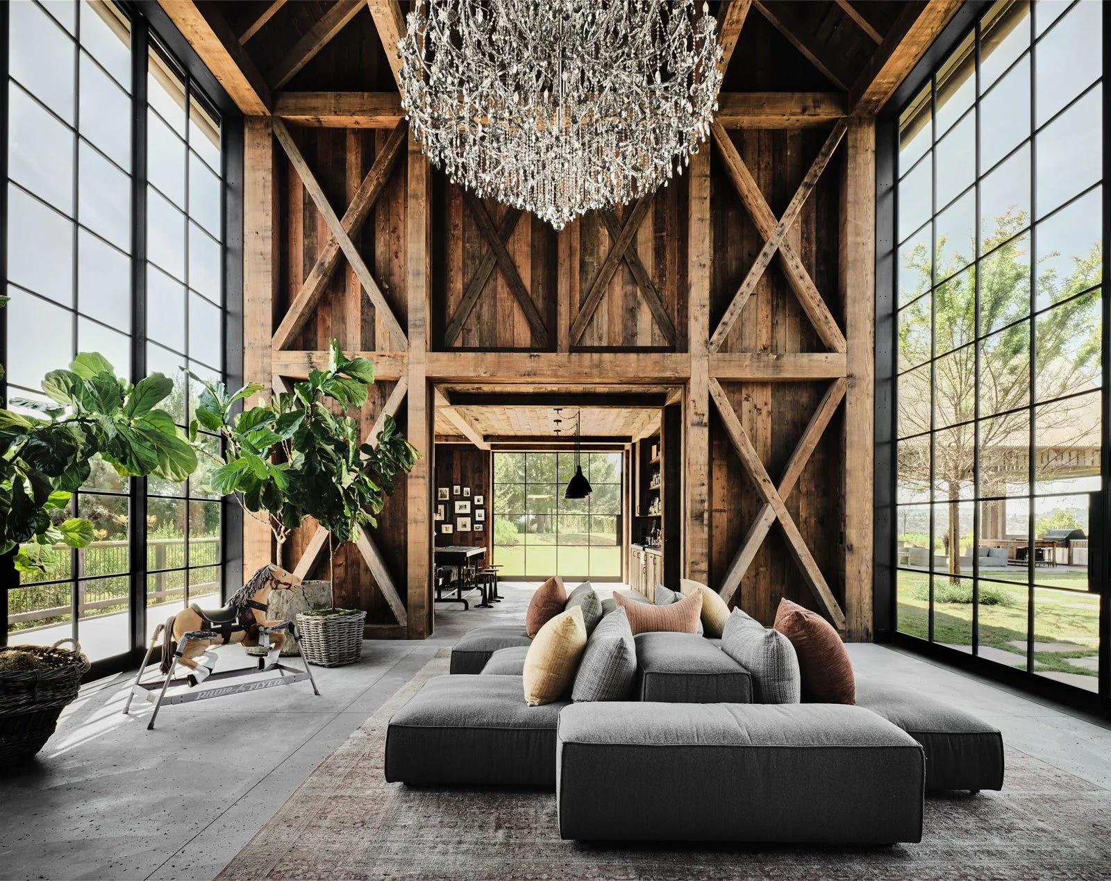 Mila Kunis & Ashton Kutcher Residence. Interior of a rustic living room with large grid windows, wooden barn-style walls, a modern gray sectional sofa with many colorful pillows, a vintage rocking horse, and a chandelier hanging from the ceiling.