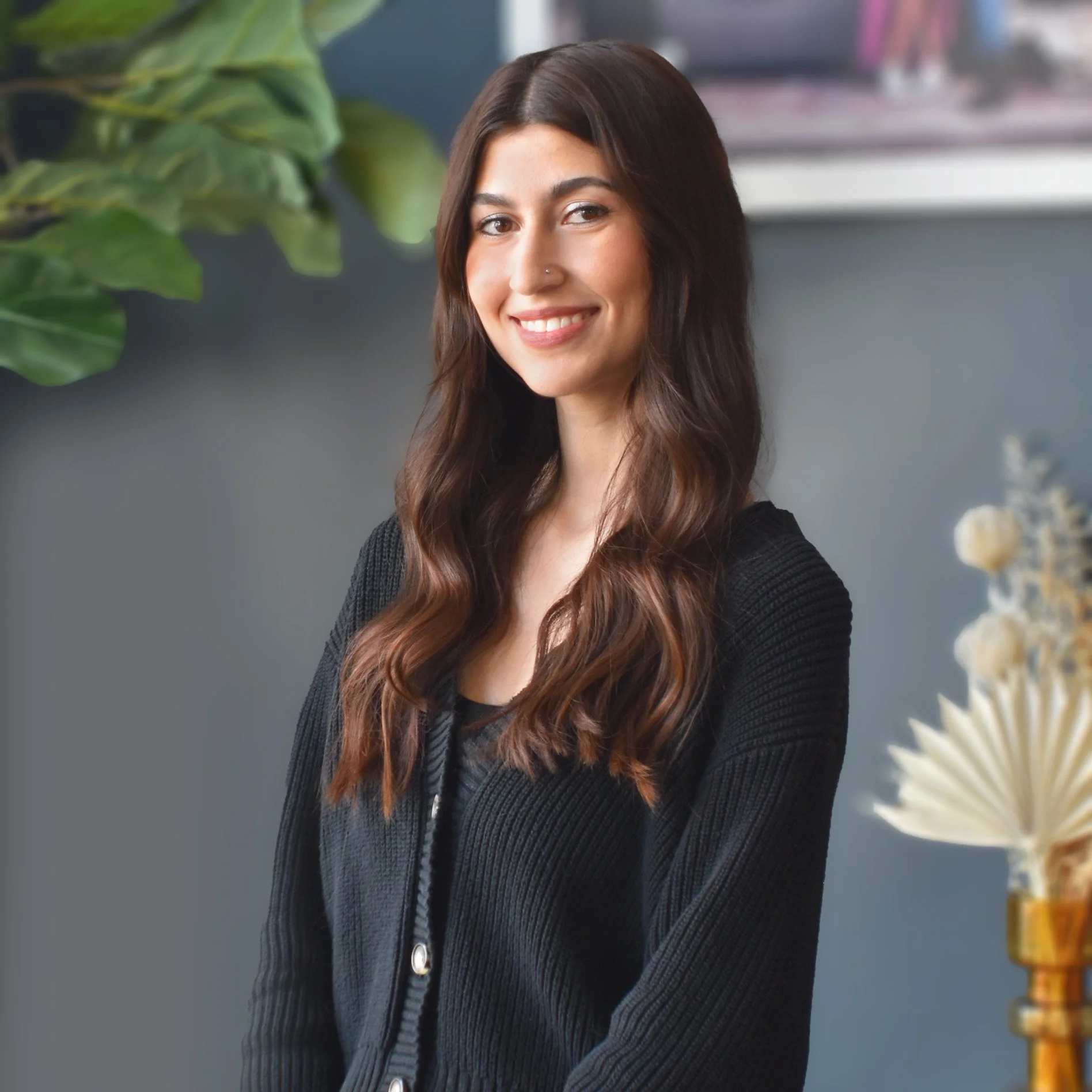 A young woman with long brown hair, wearing a black blazer and jewelry, standing indoors with a neutral expression. There are plants and a vase with dried decorative leaves on a table nearby. A large poster with an image of a couple sitting on a couch is in the background.