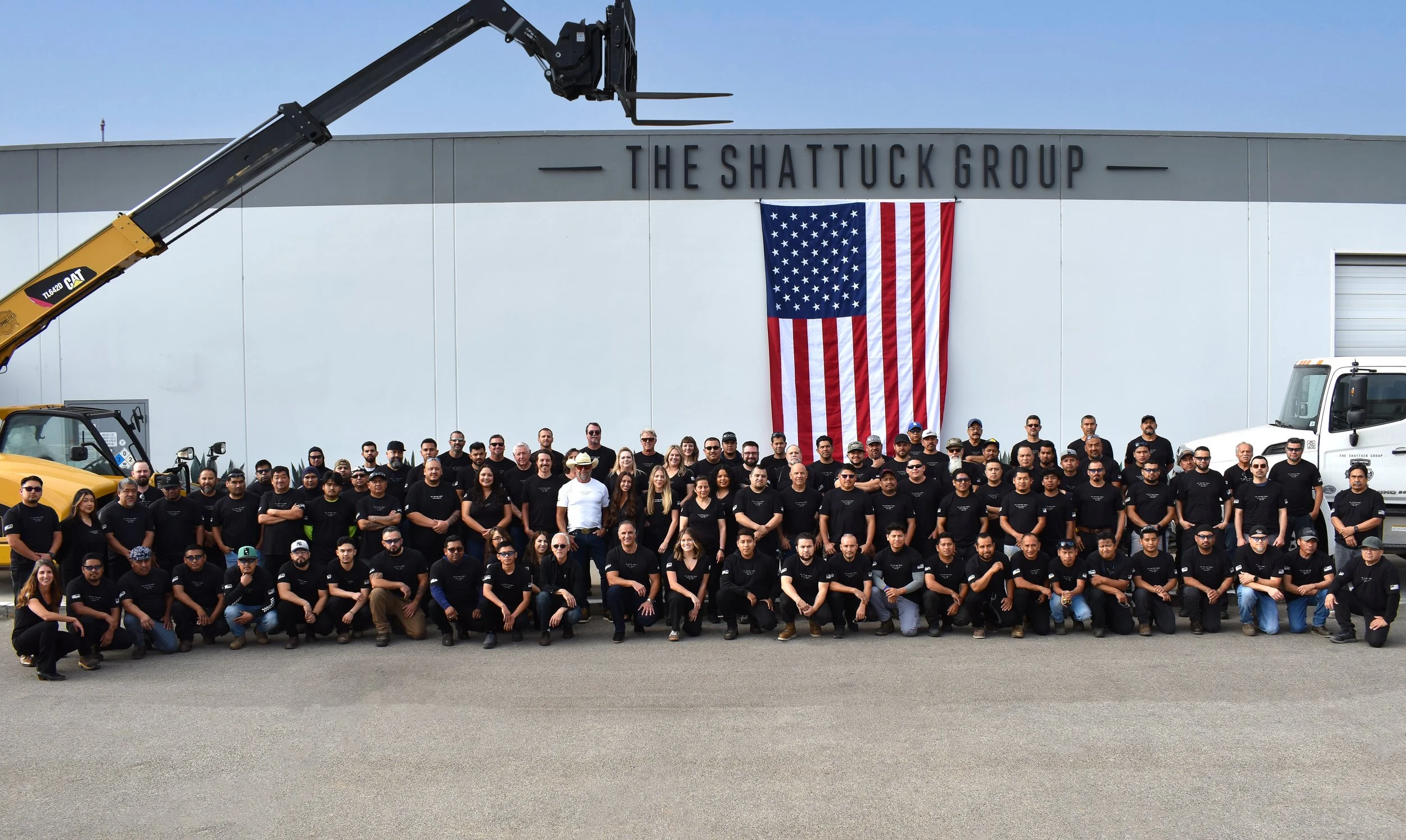 Group of people posing in front of a building with a large American flag and the sign 'The Shattuck Group' on the wall.