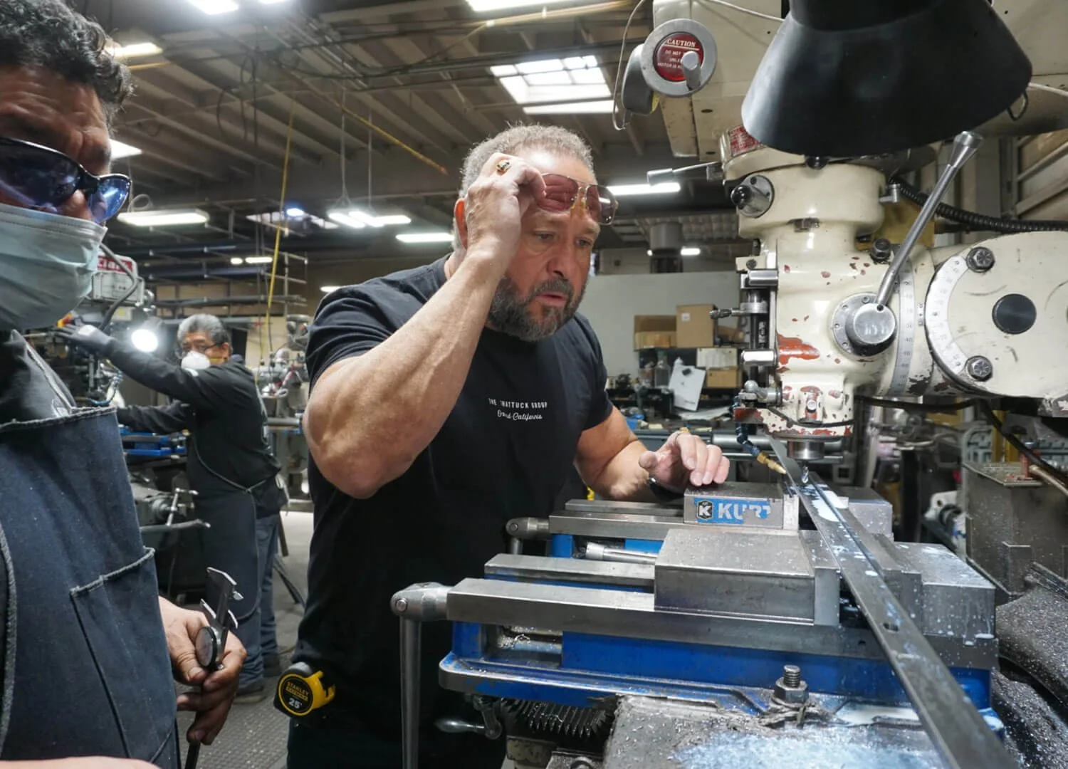 A man with a beard and black shirt working on a machine in a workshop, holding safety glasses to his forehead, with two other men wearing masks and safety glasses nearby.