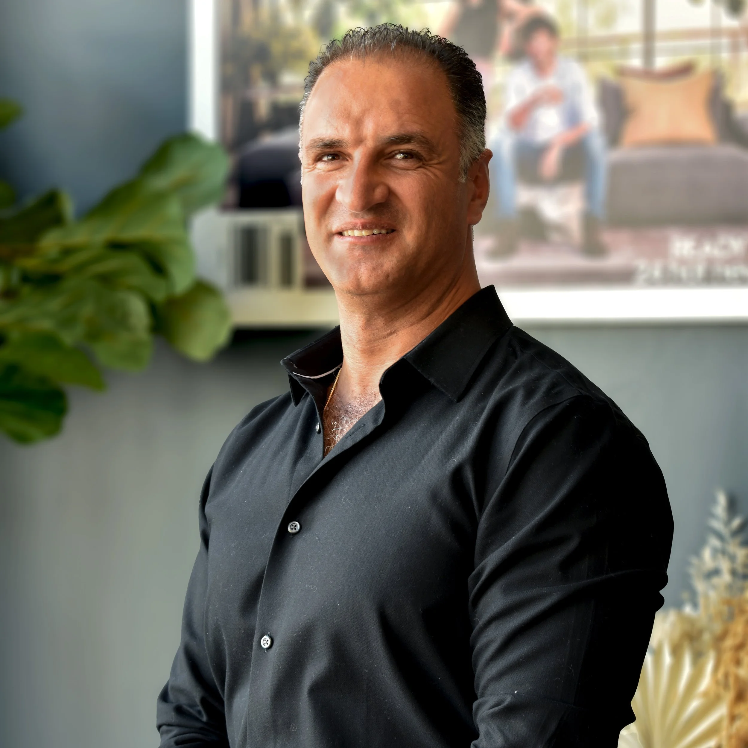 A man with short dark hair wearing a black button-up shirt, standing indoors with a potted plant and blurred background of a television and a person sitting on a couch.