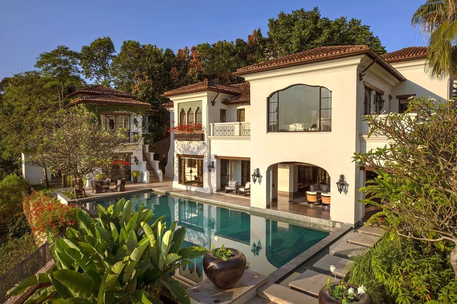 A luxurious multi-story house with white walls and brown tiled roofs, a swimming pool in the foreground, surrounded by greenery, plants, and trees under a clear blue sky.
