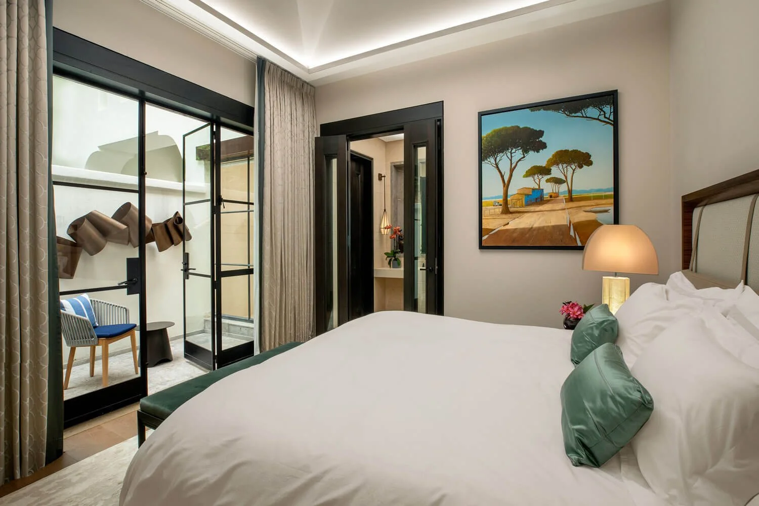 Bedroom with large bed, green pillows, bedside lamp, framed landscape painting, sliding glass door with curtains leading to a small balcony with chairs, and a view of a bathroom with a sink.