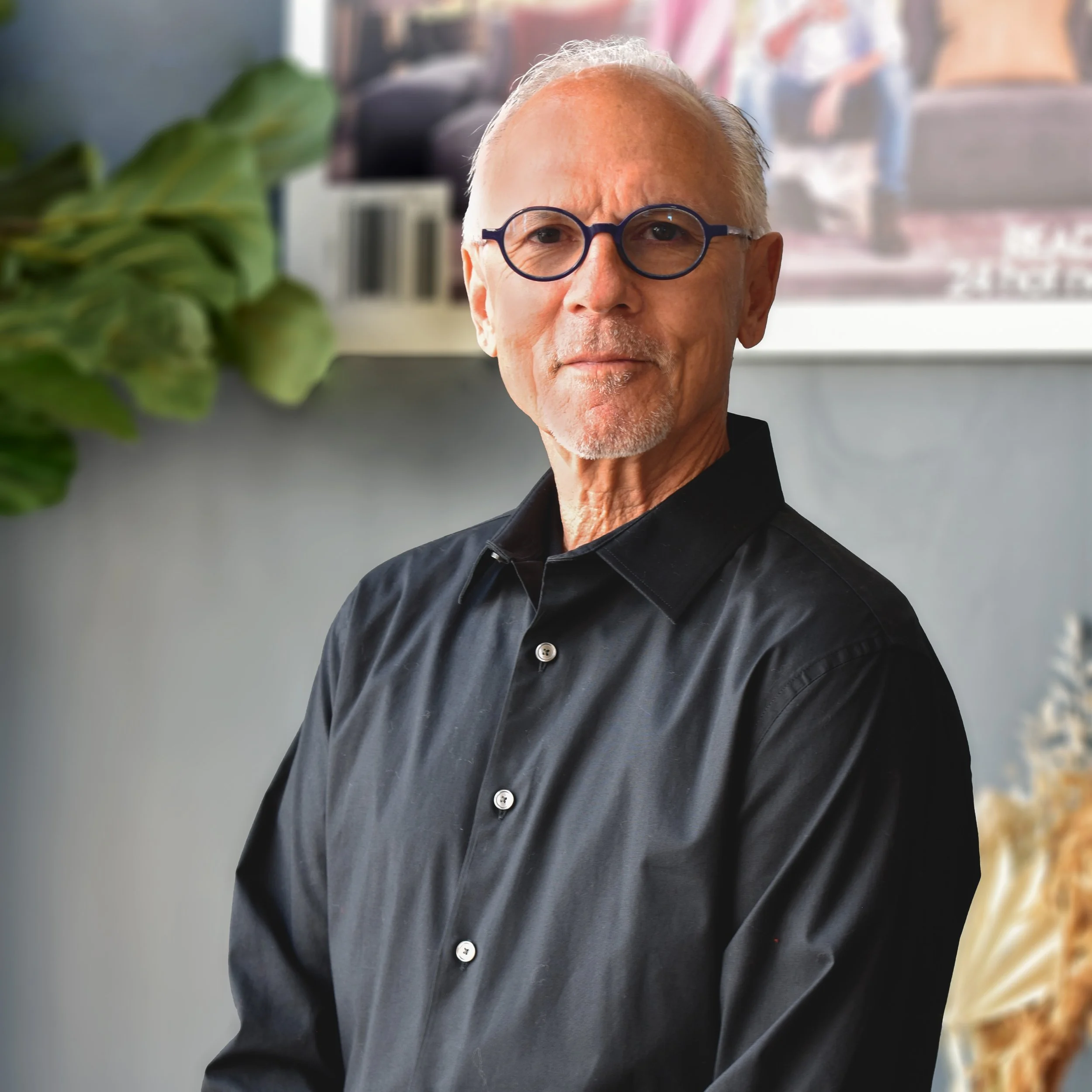 An older man with glasses and a goatee, wearing a black button-up shirt, sitting indoors with a blurred background that includes a plant and wall decor.