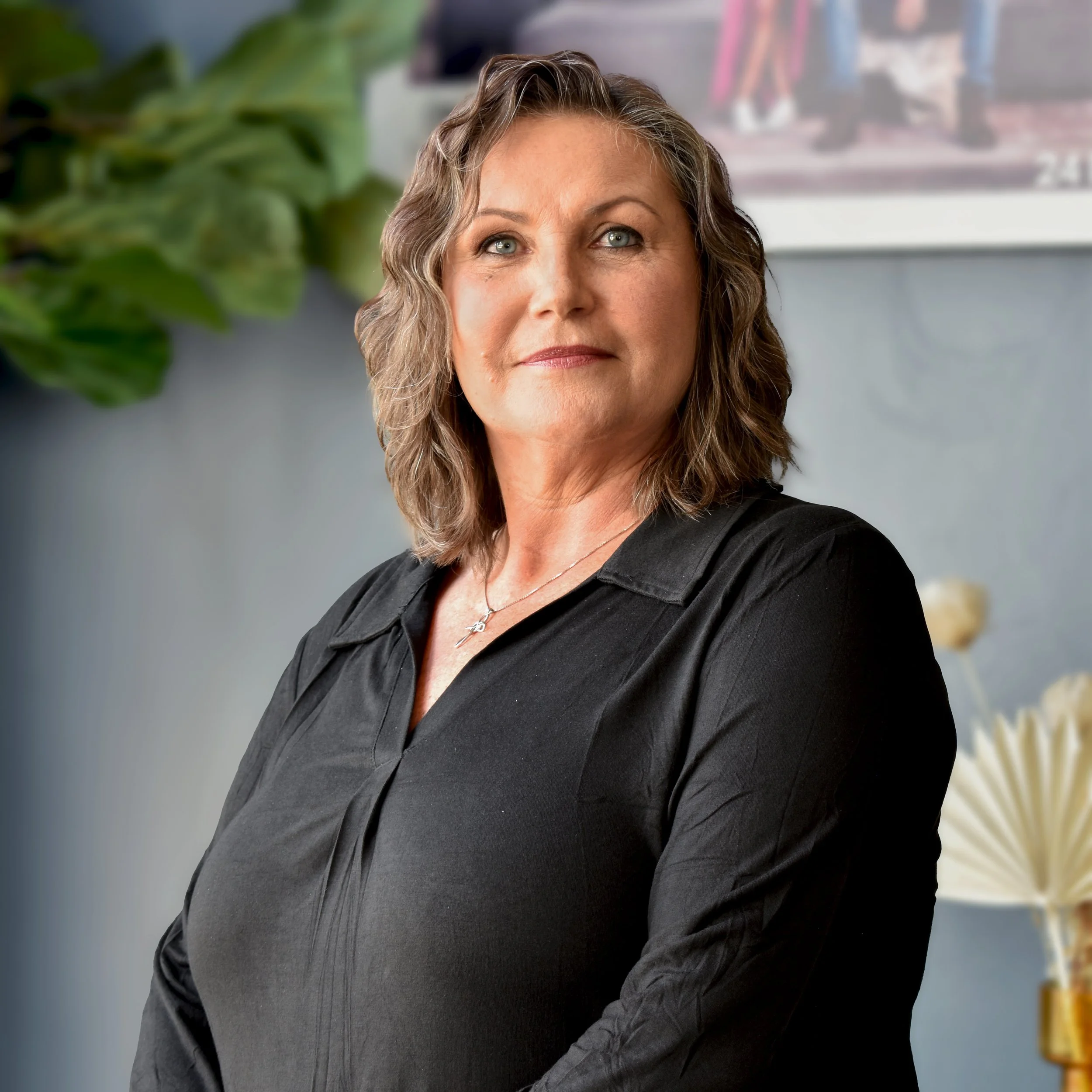 A middle-aged woman with wavy, shoulder-length hair, wearing a black shirt, standing indoors with plants and decorative items in the background.