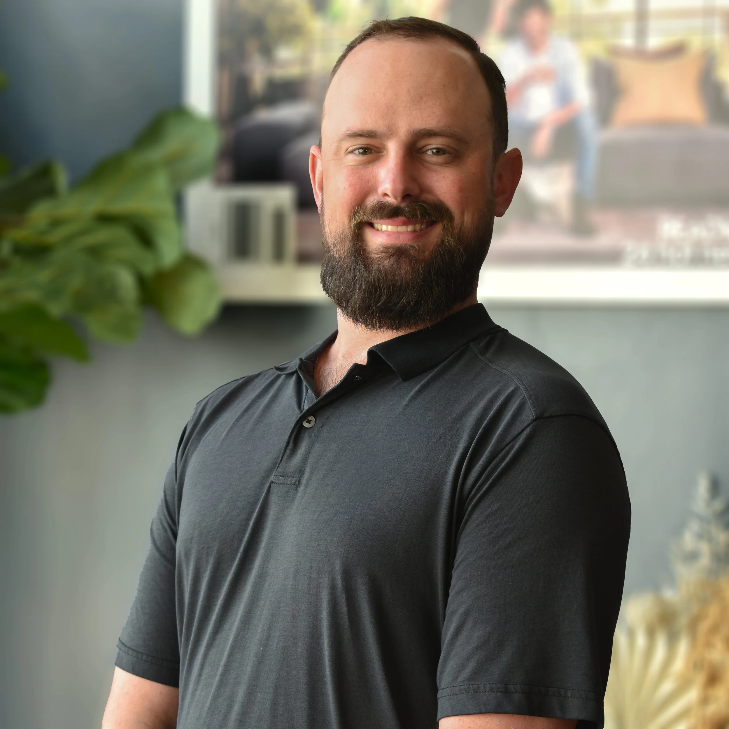 Portrait of a smiling man with a beard and short hair, wearing a black polo shirt, indoors with a blurred background.