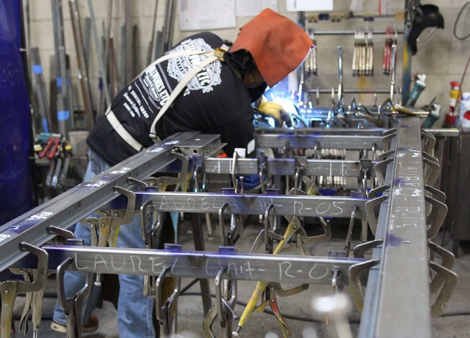 A welder wearing a protective hood and gloves working on a metal structure in a workshop. The workspace contains various metal tools and frames, with handwritten labels on the metal pieces.