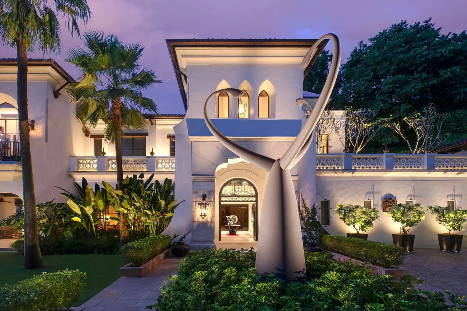 Luxury villa with Mediterranean architecture, landscape with palm trees and plants, modern sculpture in front, and warm exterior lighting at dusk.