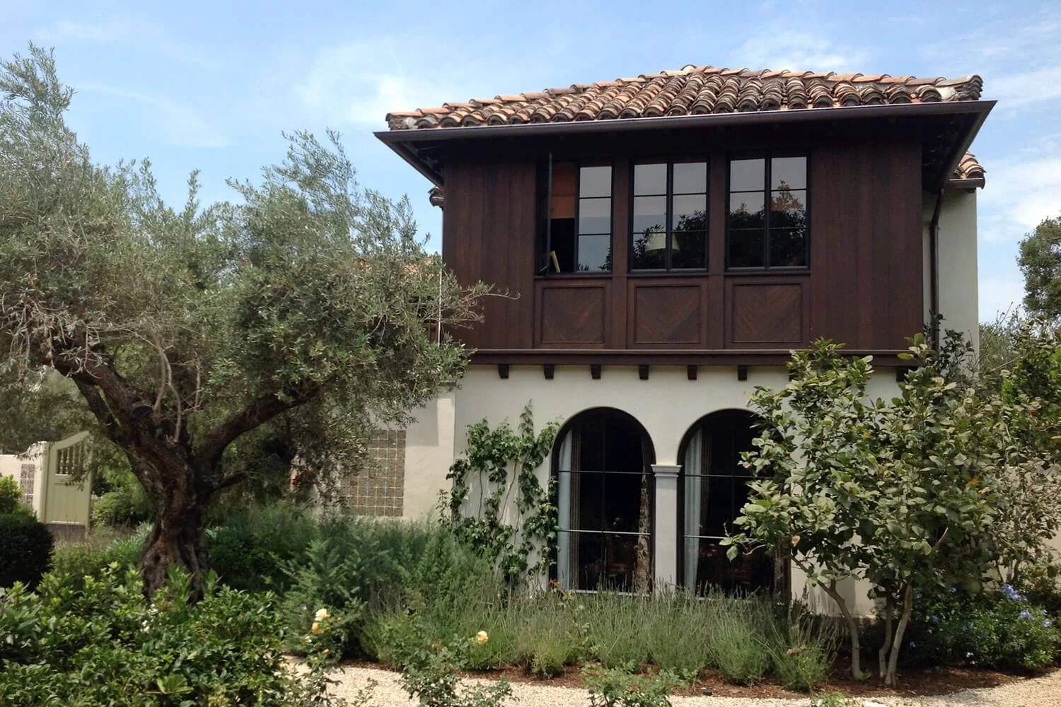 A two-story house with a white stucco lower level and a dark wooden upper level. The house has a terracotta tile roof, large black-framed windows, and arched windows on the ground floor. There is lush greenery, including a large tree and bushes, surr