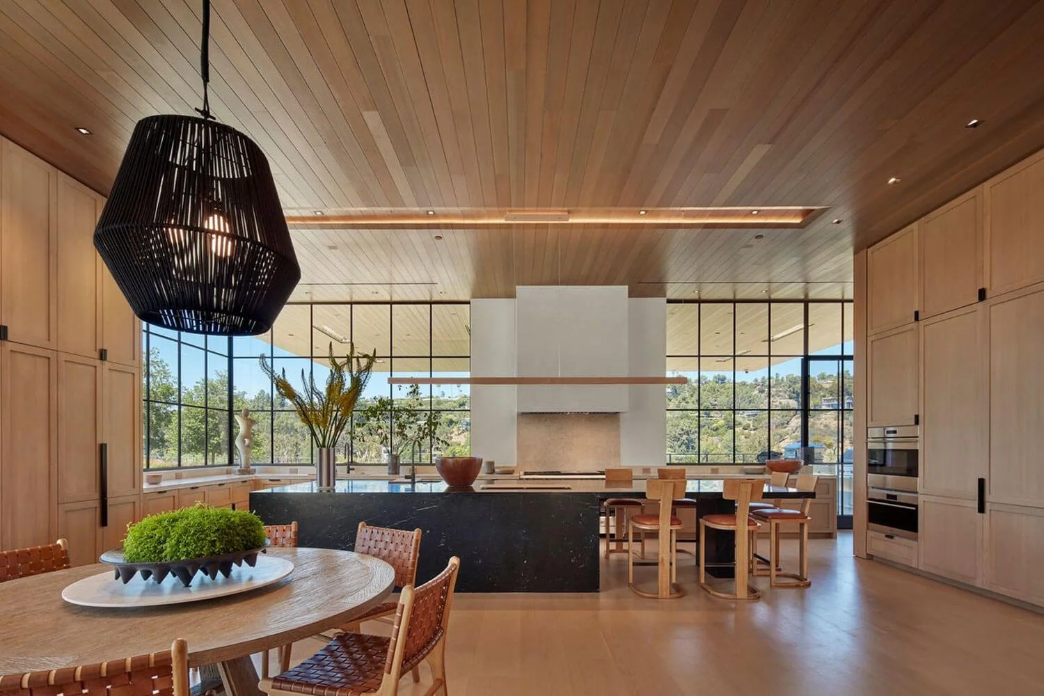 Modern kitchen with wooden cabinets, a black island, a white fireplace, large windows showing a scenic outdoor view, and a round dining table with wicker chairs.