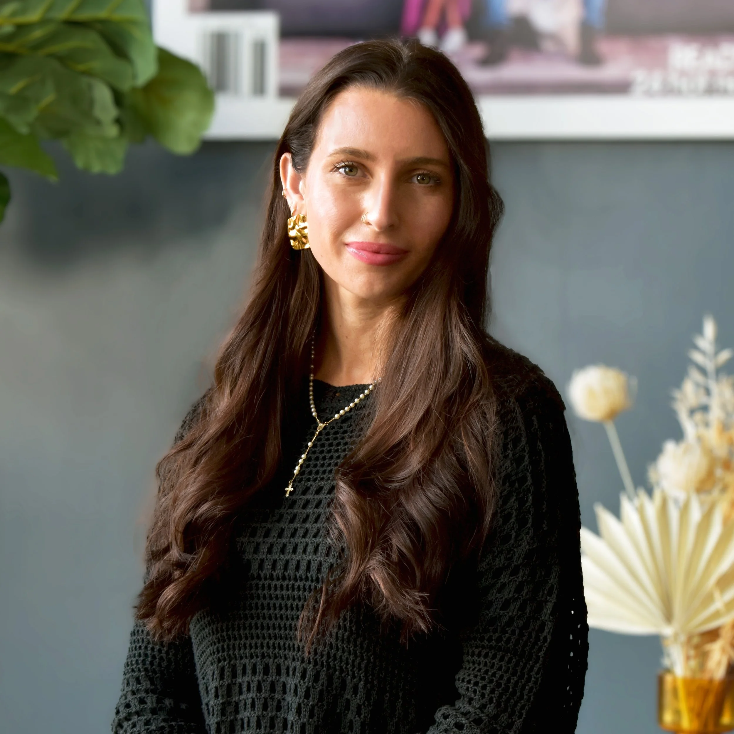 A woman with long brown hair, wearing a black shirt, standing indoors against a gray wall with decorative plants in the background.