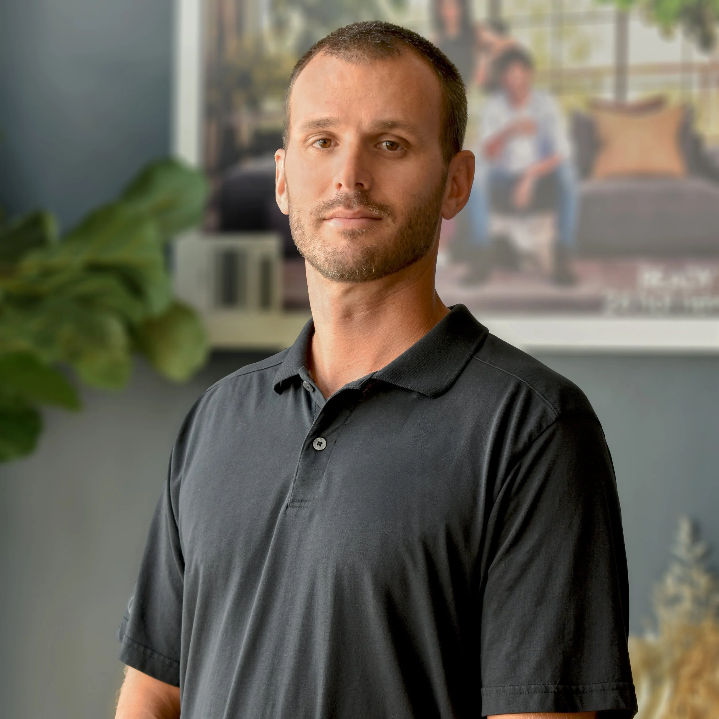 Portrait of a man wearing a black polo shirt, standing indoors with a blurred background of a person sitting on a couch.