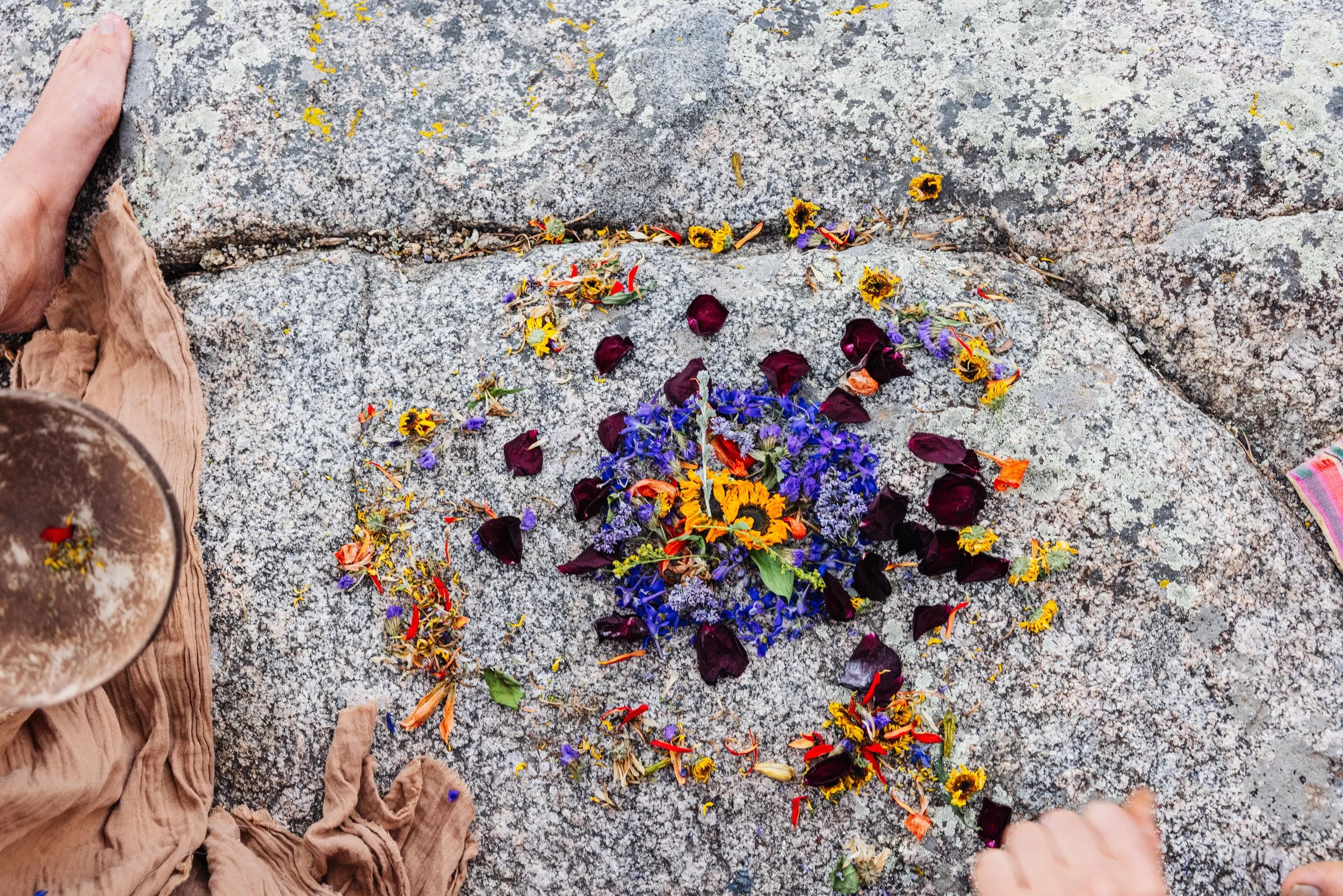 Colorful flower arrangement on a stone surface, with a person on the left wearing a beige skirt and holding a brown item, and a person on the right with a hand pointing toward the flowers.