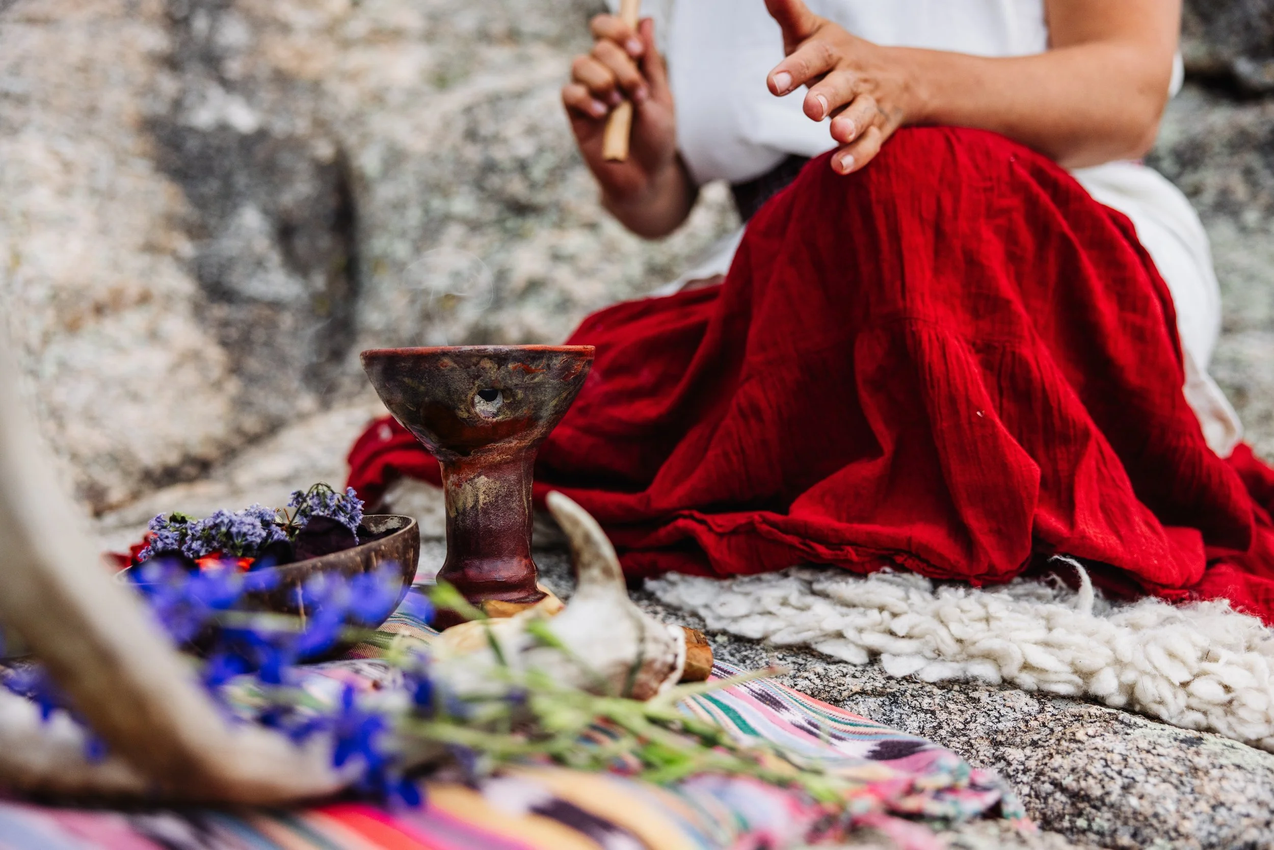 Person sitting on a blanket with herbs, flowers, a carved wooden bowl, and a ceramic chalice, dressed in a white shirt and a bright red skirt, holding a small wooden flute, outdoors on a rocky surface.