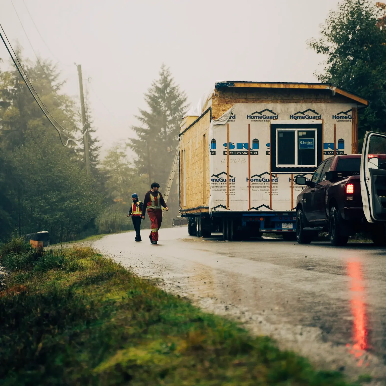 Construction workers walking near a house under construction on a foggy road, with trees and a utility pole in the background.