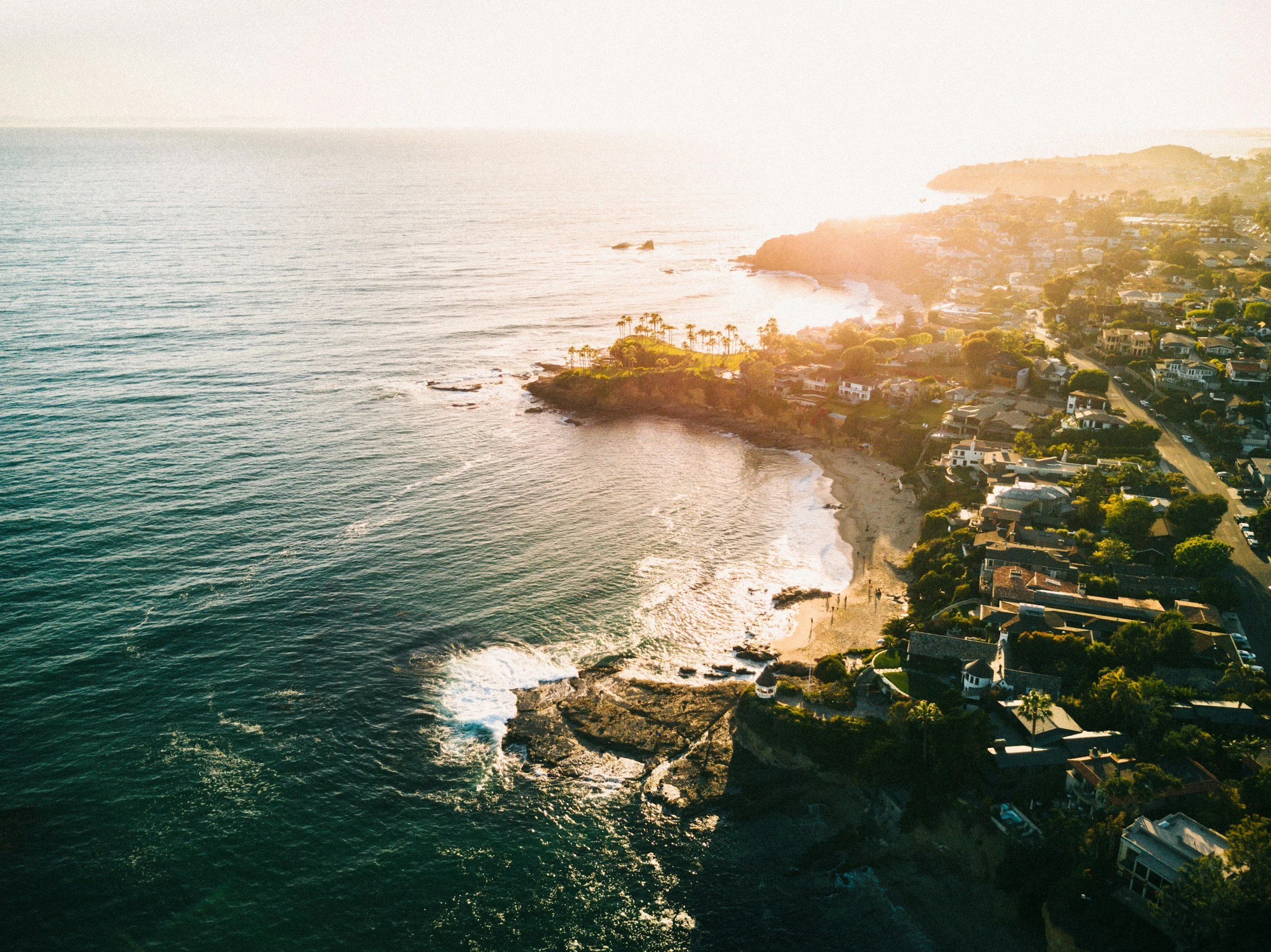 Aerial view of a coastal town at sunset, showing houses along the shoreline, a small beach, and the ocean extending to the horizon.