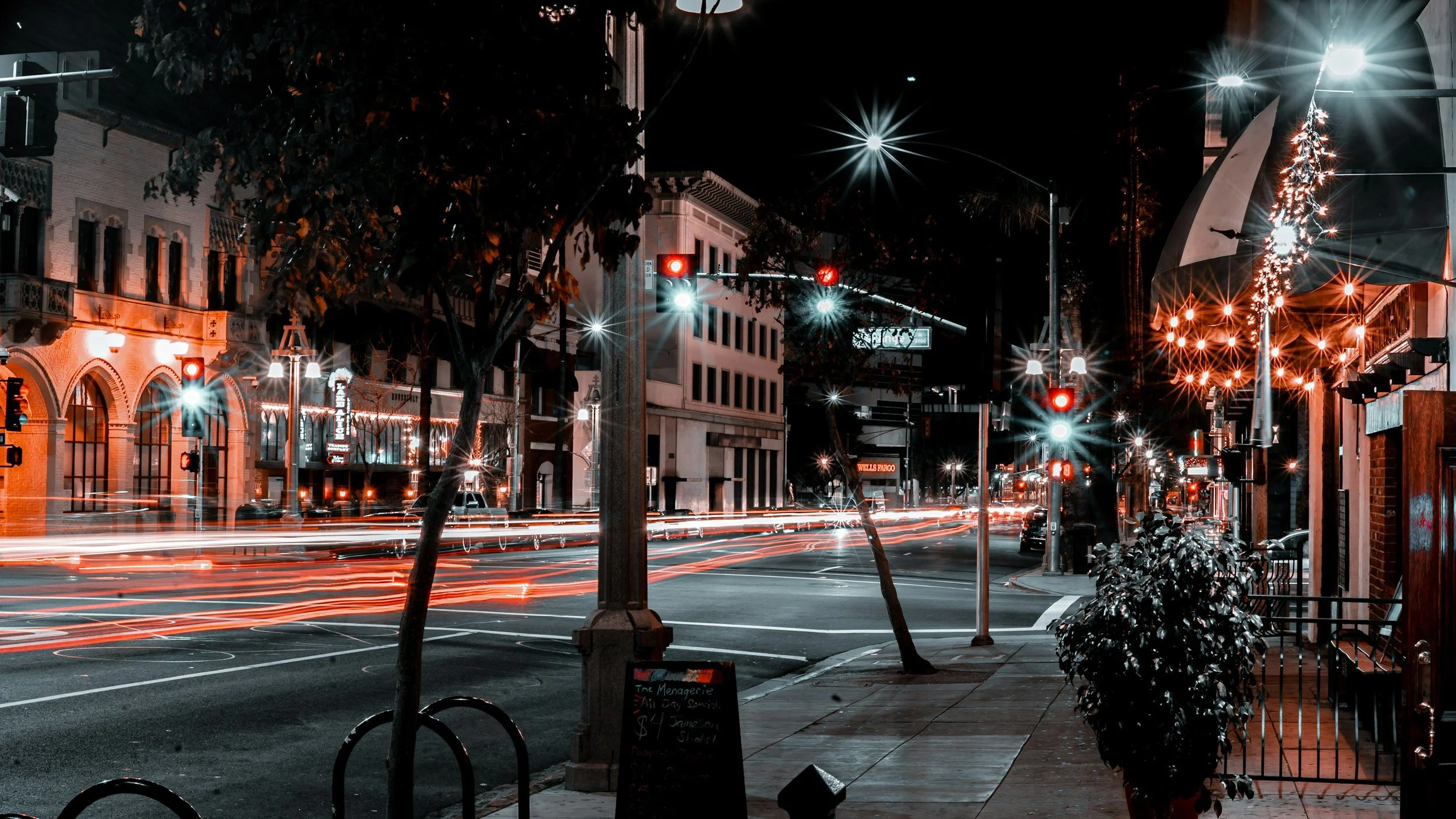 Nighttime city street scene with traffic lights, light trails from passing cars, buildings with illuminated signs, and sidewalk plants.