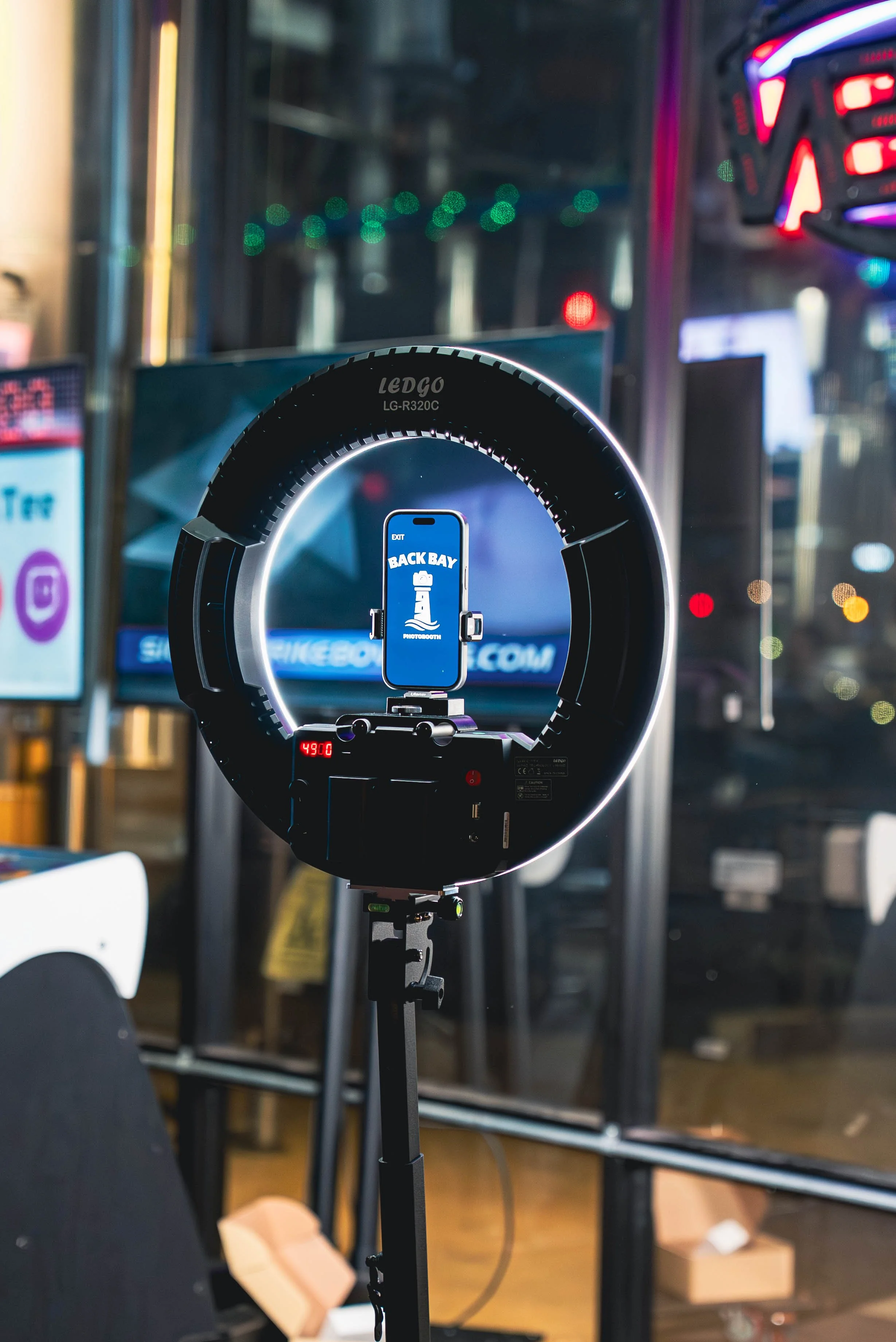Ring light setup on tripod with a smartphone displaying 'Back Bay Photobooth' and a lighthouse logo.