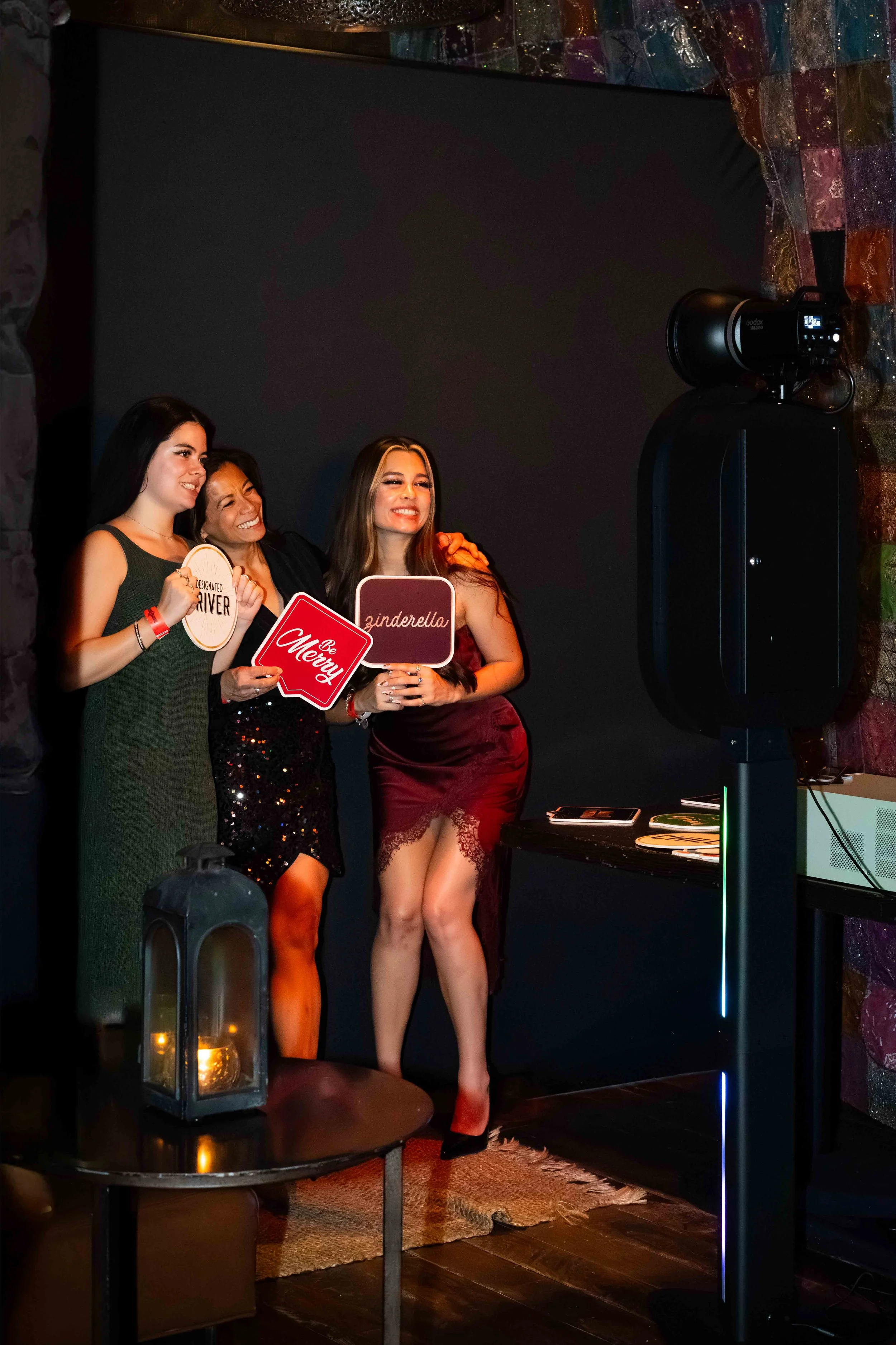 Three women dressed in elegant evening wear taking a photo at a party with photo props, in front of a black background with colorful decorations.