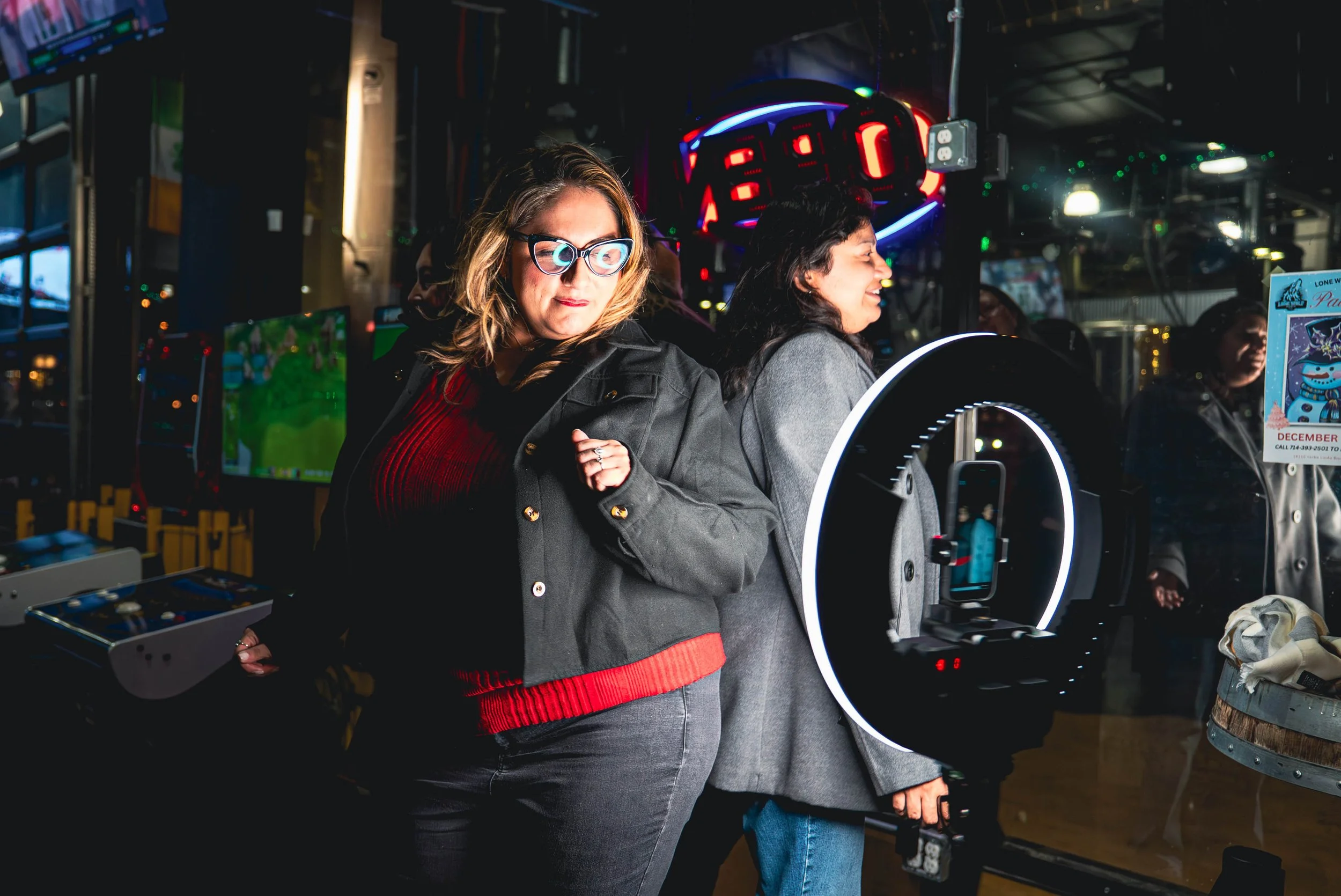 Two women standing in an indoor arcade or entertainment center next to a ring light with a smartphone attached, with arcade machines and a scoreboard in the background.