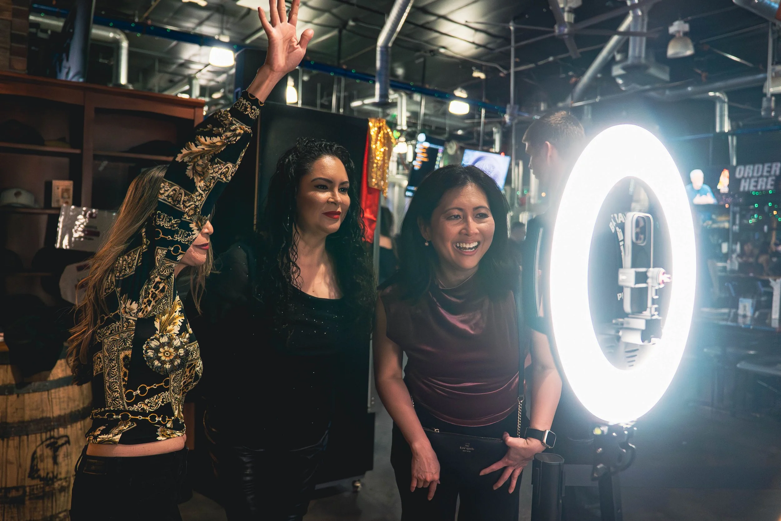 Three women and one man looking at a camera with ring light in a bar or restaurant, with one woman smiling and the others observing.