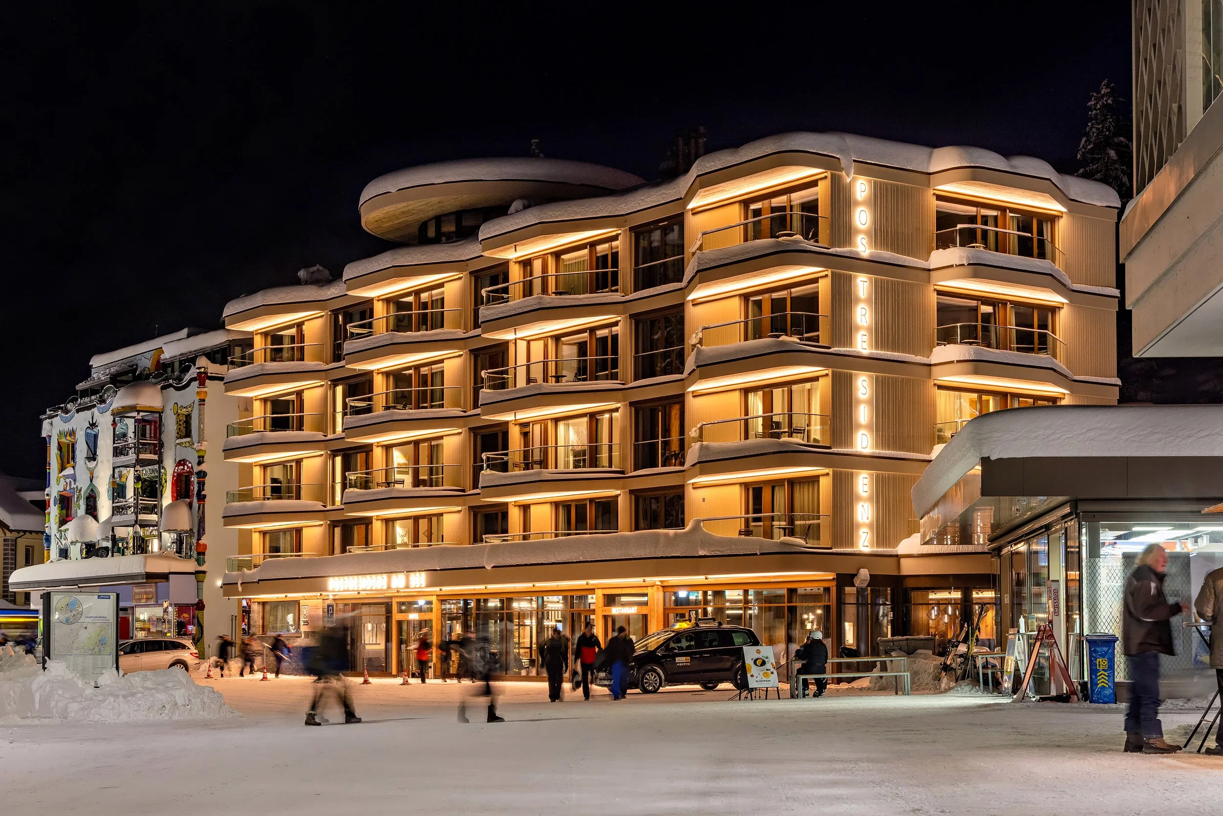Mehrstöckiges Hotel in winterlicher Umgebung bei Nacht, mit beleuchteten Balkonen, Schnee auf den Dächern und Schneemassen am Boden, mit Menschen im Vordergrund.