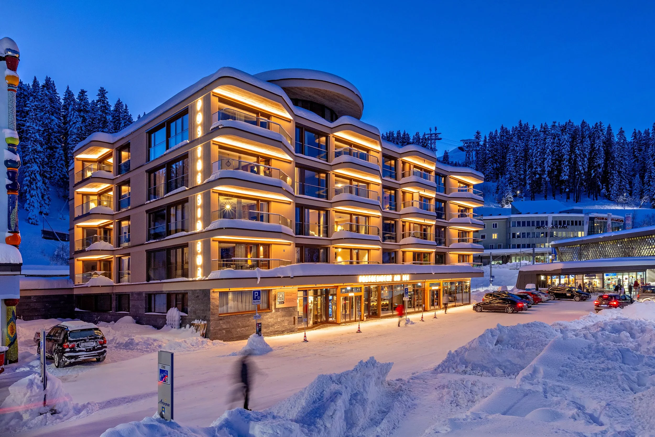 Ein beleuchtetes Hotel in einer verschneiten Bergregion bei Dämmerung, mit Parkplätzen und Schneehaufen vor dem Gebäude. Im Hintergrund sind schneebedeckte Tannen und Skilifte sichtbar.