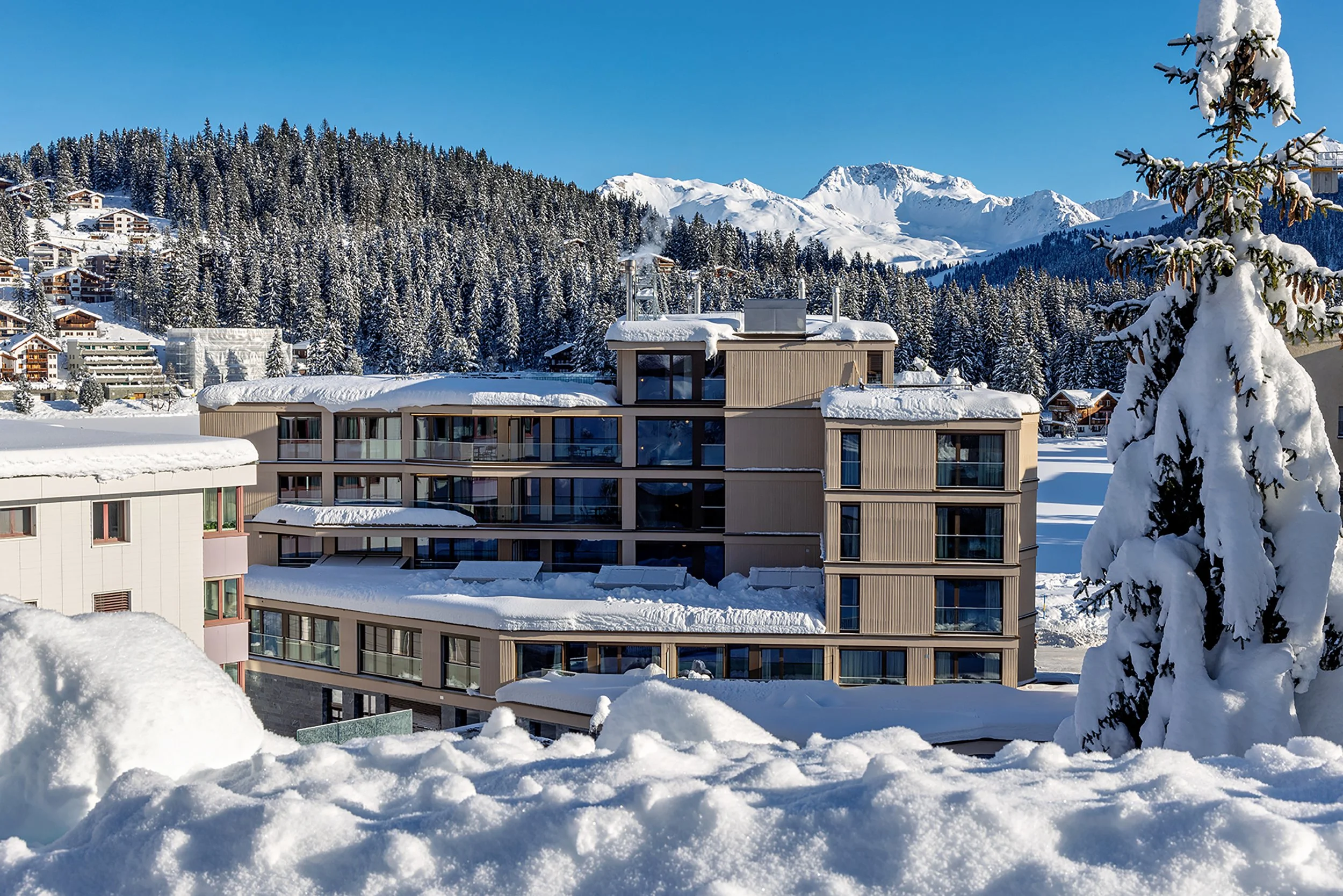 Moderne Gebäuden in einer verschneiten Bergregion mit schneebedeckten Bäumen und Bergen im Hintergrund, bei blauem Himmel.