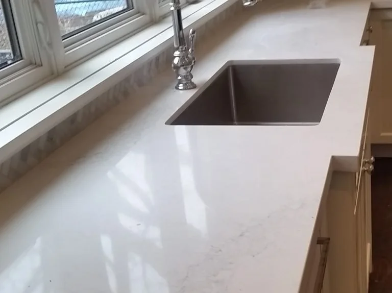 A white marble kitchen countertop with a stainless steel sink and a chrome faucet, next to a window.