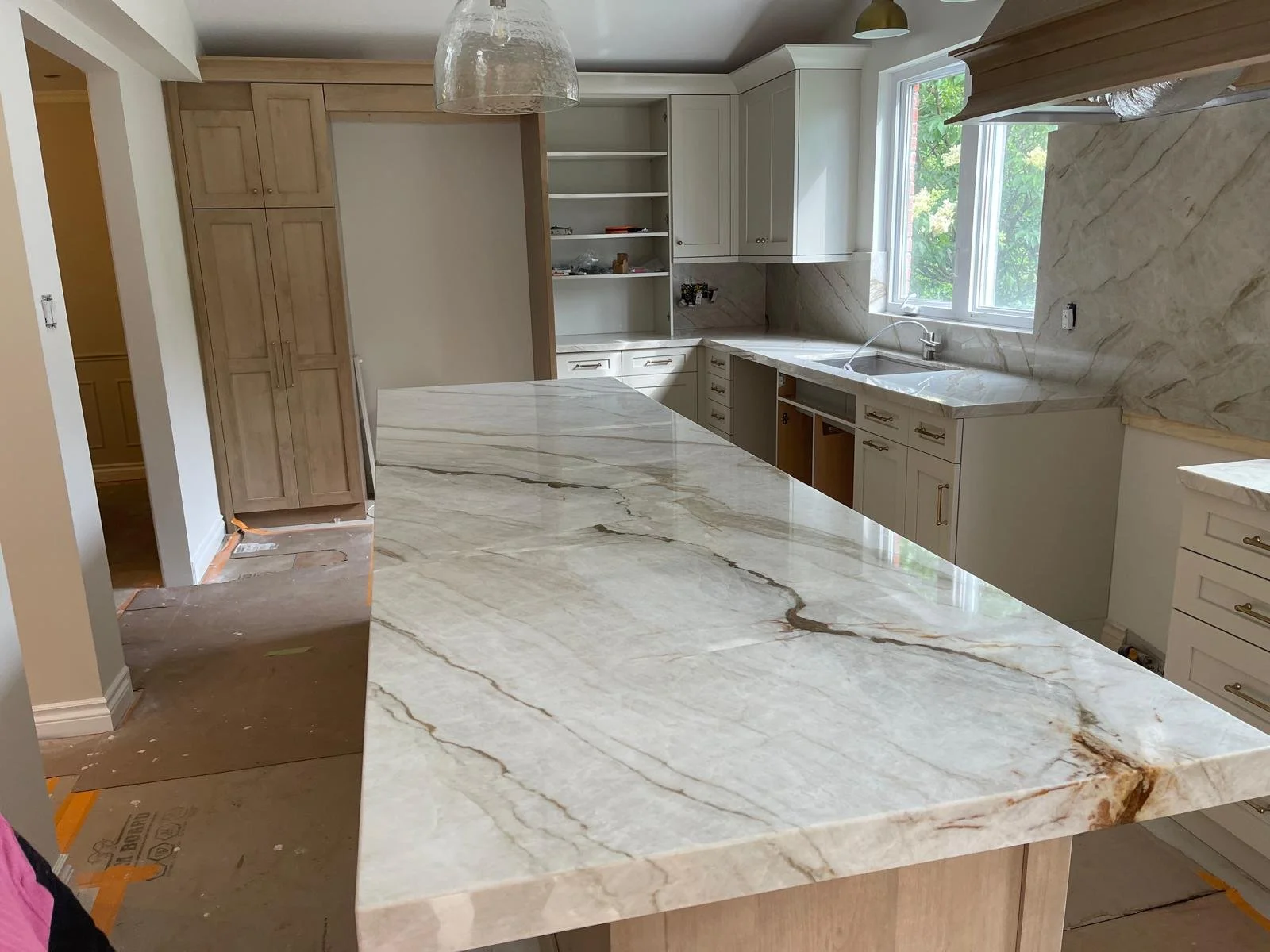 Kitchen with an island, white cabinetry, marble countertops and backsplash, a sink under a window, and partially open cabinets, in a home under renovation.