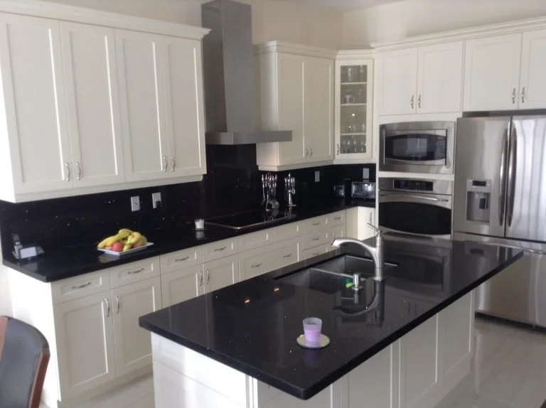 Modern kitchen with white cabinets, black countertops, stainless steel appliances, including a refrigerator, microwave, and oven. There is a kitchen island with a sink, a fruit bowl with bananas, and small decorative items on the counters.