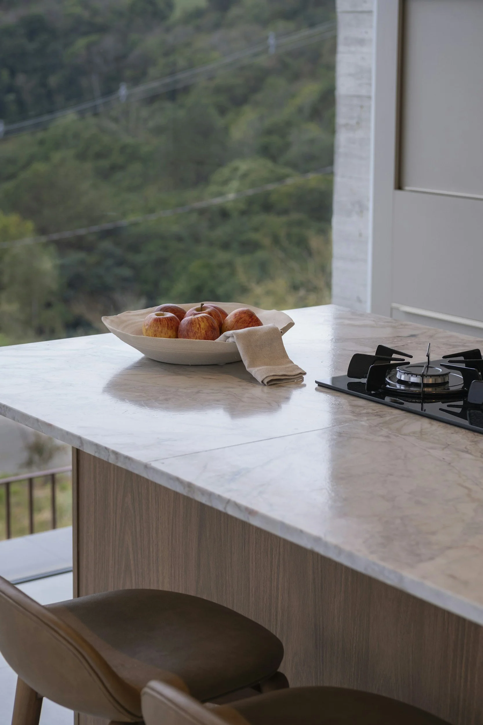 A kitchen countertop with a white bowl of apples, a beige napkin, and a stovetop, with a scenic view of green hills outside the window.