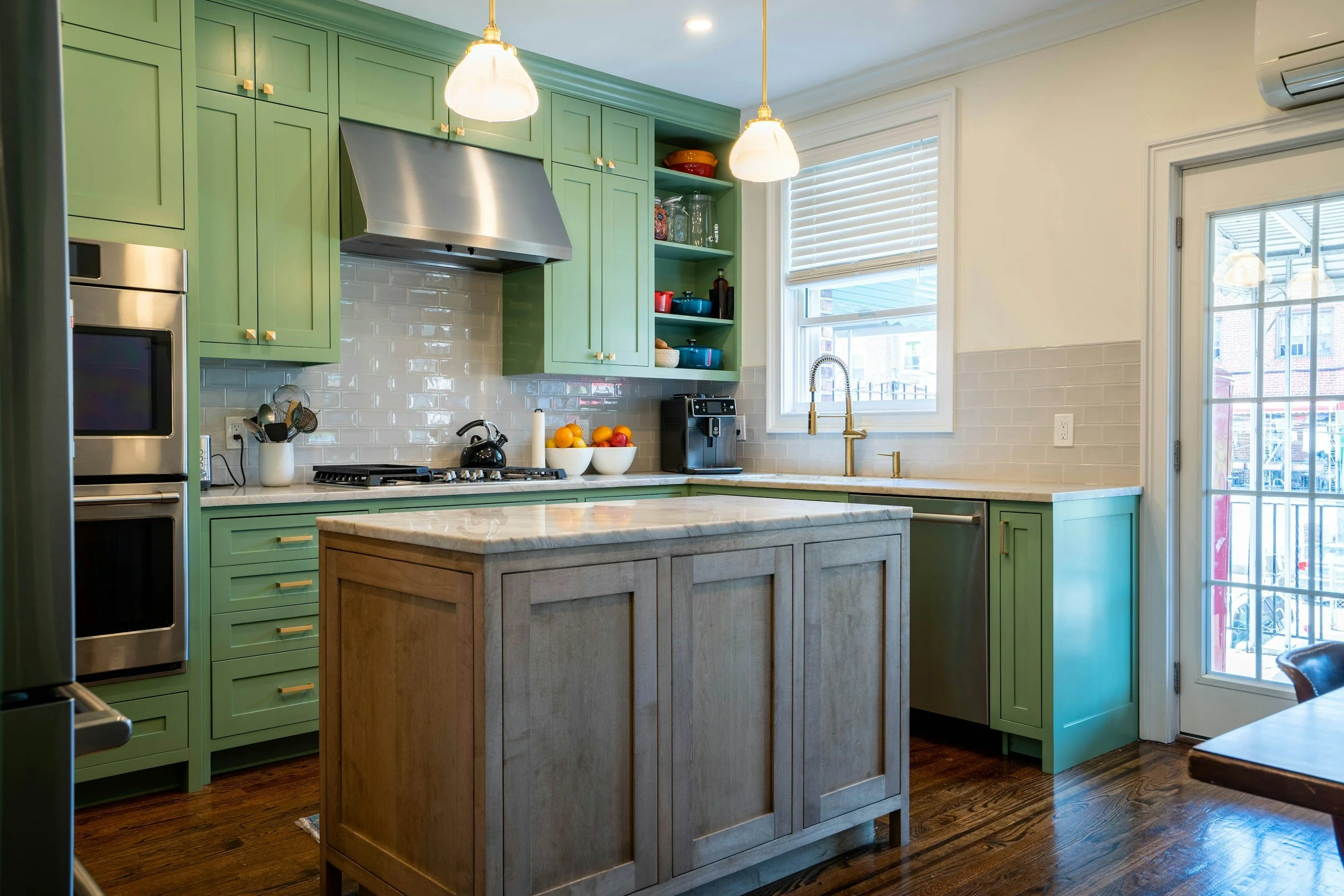 A modern kitchen with green cabinets, a kitchen island with a light wood finish and a marble countertop, stainless steel appliances, white subway tile backsplash, and large windows letting in natural light.