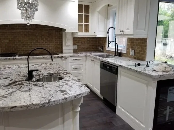 Modern kitchen with white cabinets, durable granite countertops with fancy design, a centered island with a sink and black faucet, a window over the sink, a brick backsplash, and a large window looking outside.