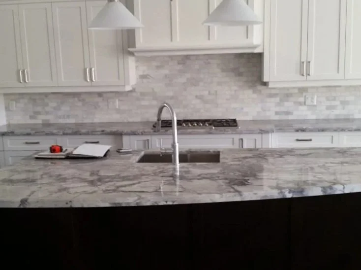 Modern kitchen with white cabinets, marble countertop island, stainless steel sink, and backsplash, pendant lights overhead.