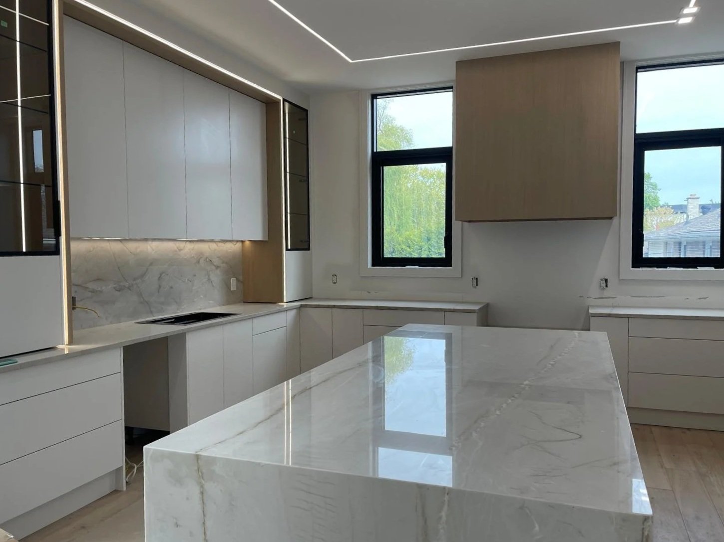 Modern kitchen with white marble countertops, beige cabinets, two large windows showing green trees outside, and minimalist lighting.