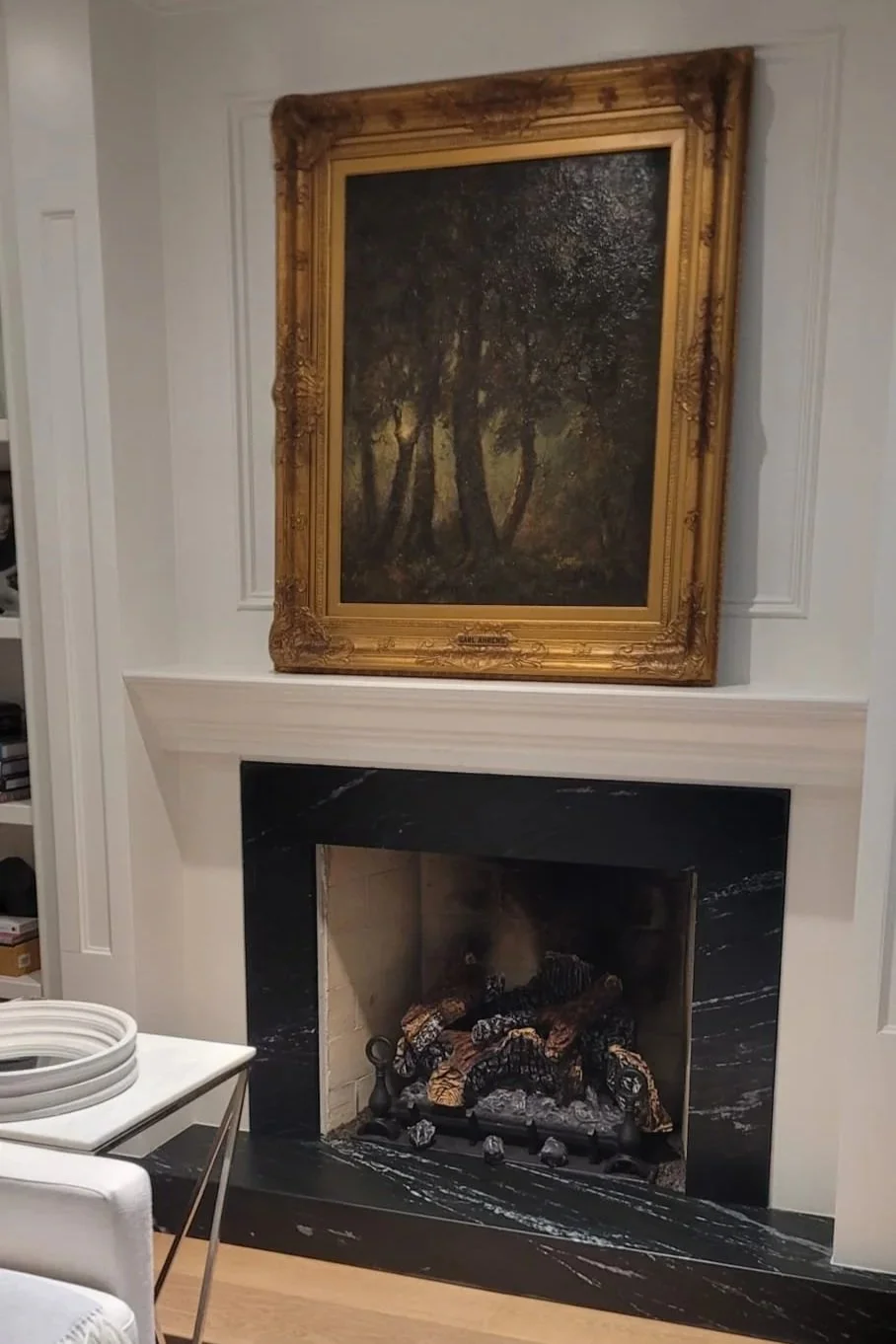 A living room fireplace with a black marble surround, a white mantle, and a painting of a forest scene hanging above it.