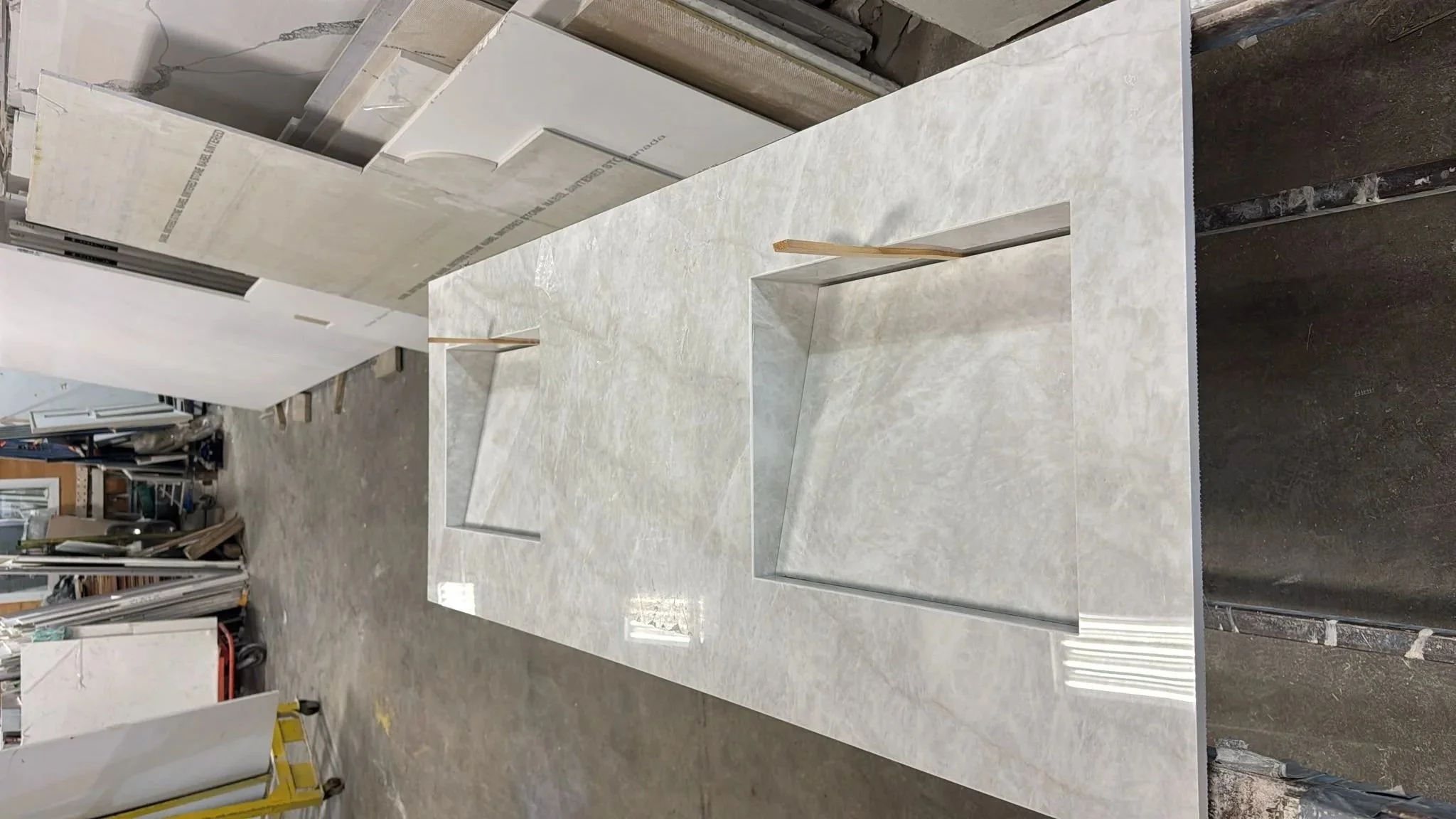 A large, rectangular, light-coloured stone or marble island with a geometric indented design in the center, placed in an unfinished workshop or warehouse setting for fabrication before installation.
