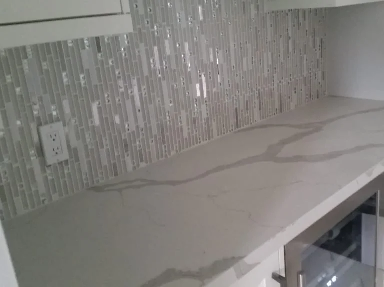 Kitchen counter with a gray marble surface and mosaic tile backsplash, electrical outlet, and part of a glass-front cabinet.