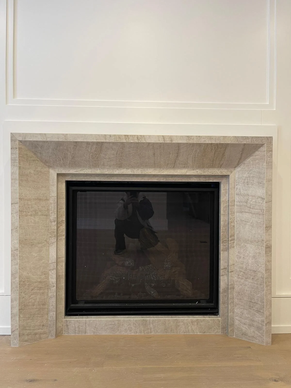 Photo of a fireplace with a black frame, surrounded by a beige stone mantel, in a room with white walls and wood flooring. Reflection of a person taking photo is visible in the fireplace glass.