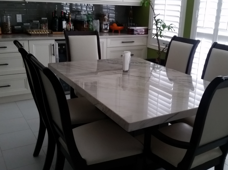 A dining room with a marble table, six chairs, a green wall, white cabinets, a window with blinds, and a countertop with bottles and kitchen items.