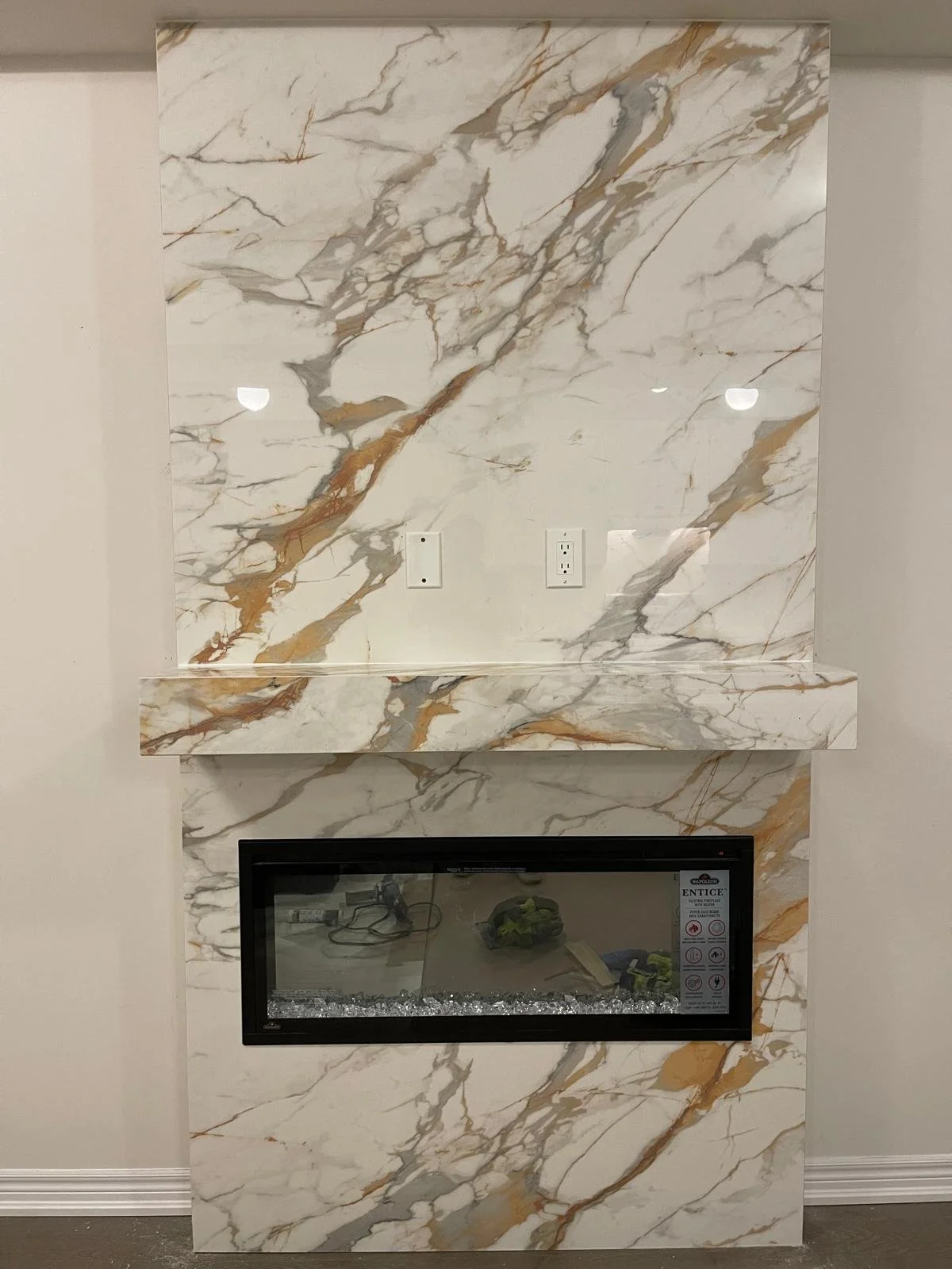 Porcelain fireplace surround in white, grey, brown with a built-in electric fire and two electrical outlets above it.