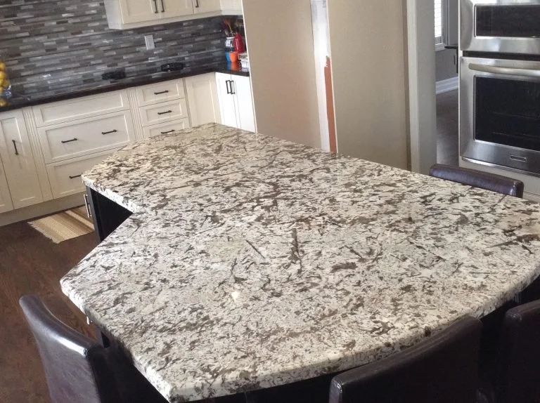 Kitchen with a large granite island countertop surrounded by dark chairs, white cabinets, and stainless steel appliances.
