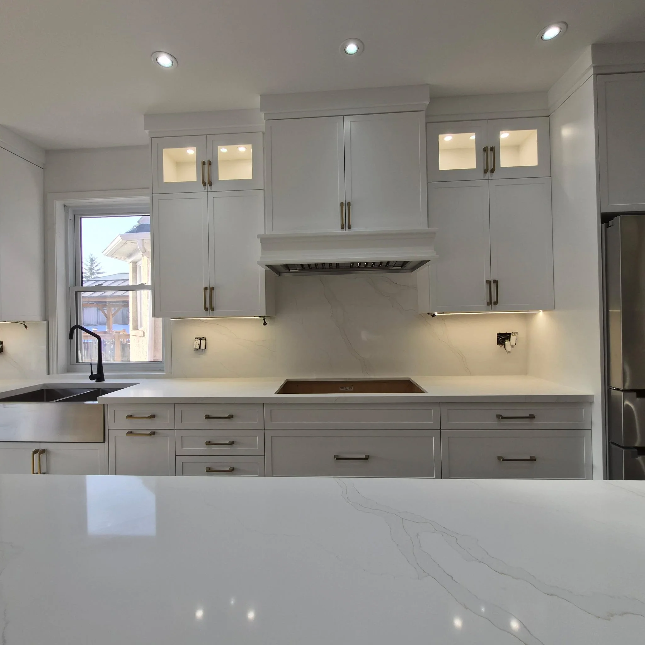 Modern white kitchen with upper and lower cabinets, a built-in range with a venthood, a double sink with a black faucet, a window, and stainless steel appliances.